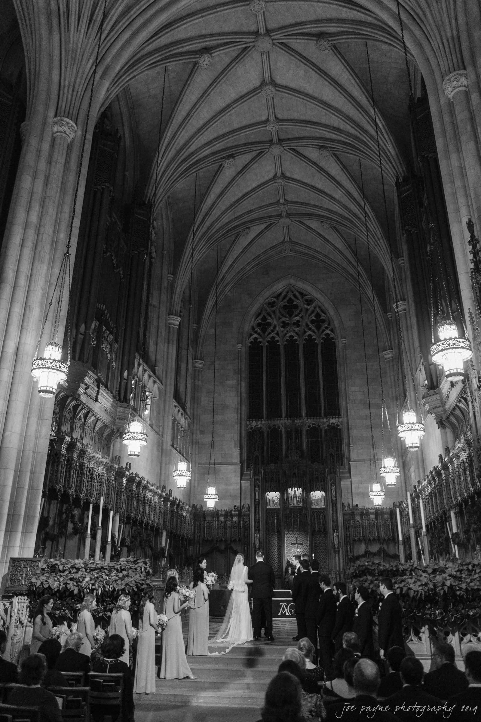 carolina inn & duke chapel wedding - kathleen & michael A black and white photo of a wedding ceremony in a grand cathedral with high vaulted ceilings. The bride and groom stand at the altar, surrounded by attendants and guests seated on either side. Chandeliers illuminate the ornate interior.