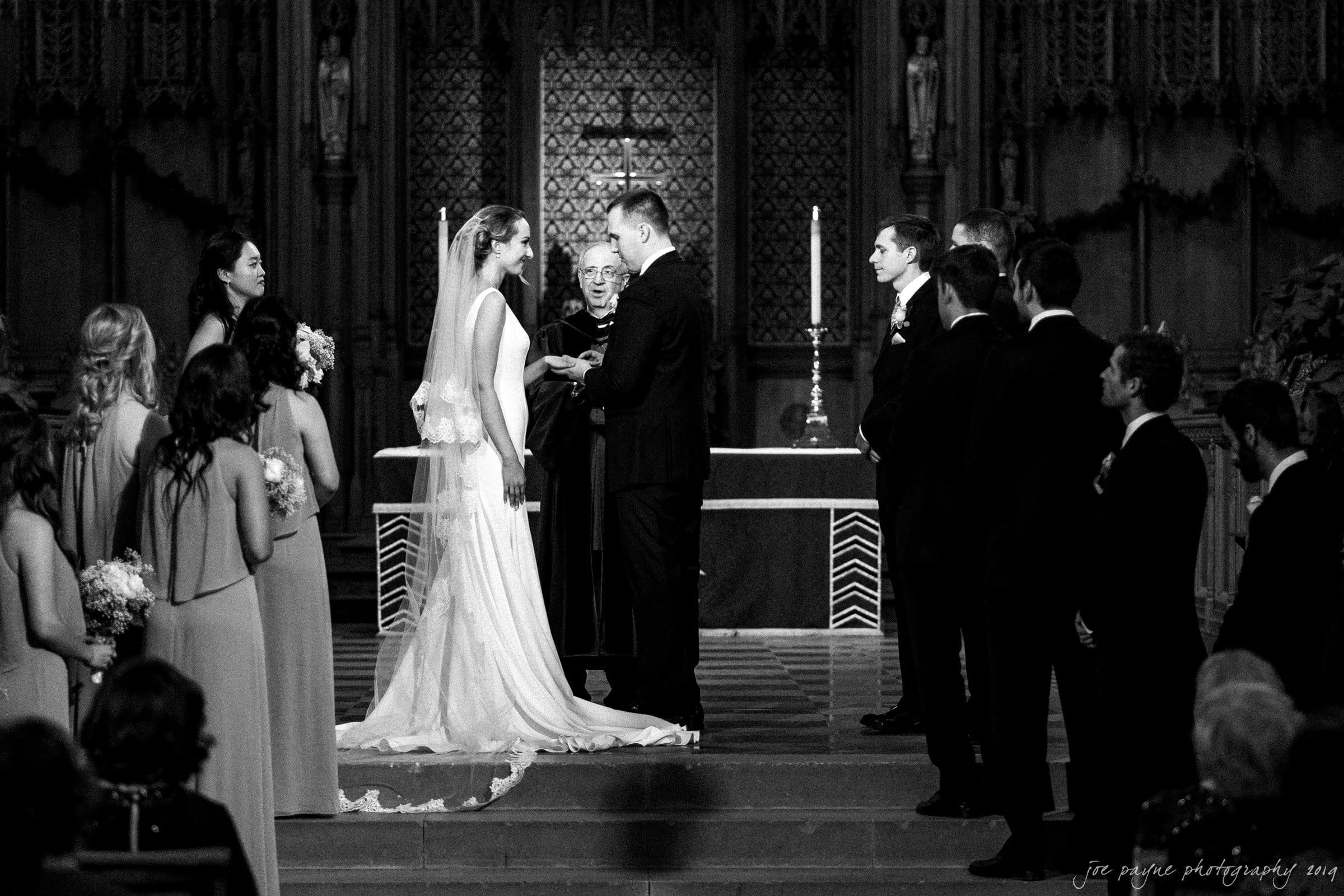 carolina inn & duke chapel wedding - kathleen & michael A black and white photo of a wedding ceremony in a church. The bride in a long gown and veil stands facing the groom in a suit. An officiant is between them. Bridesmaids and groomsmen are standing on either side. Candles and decorative patterns are in the background.
