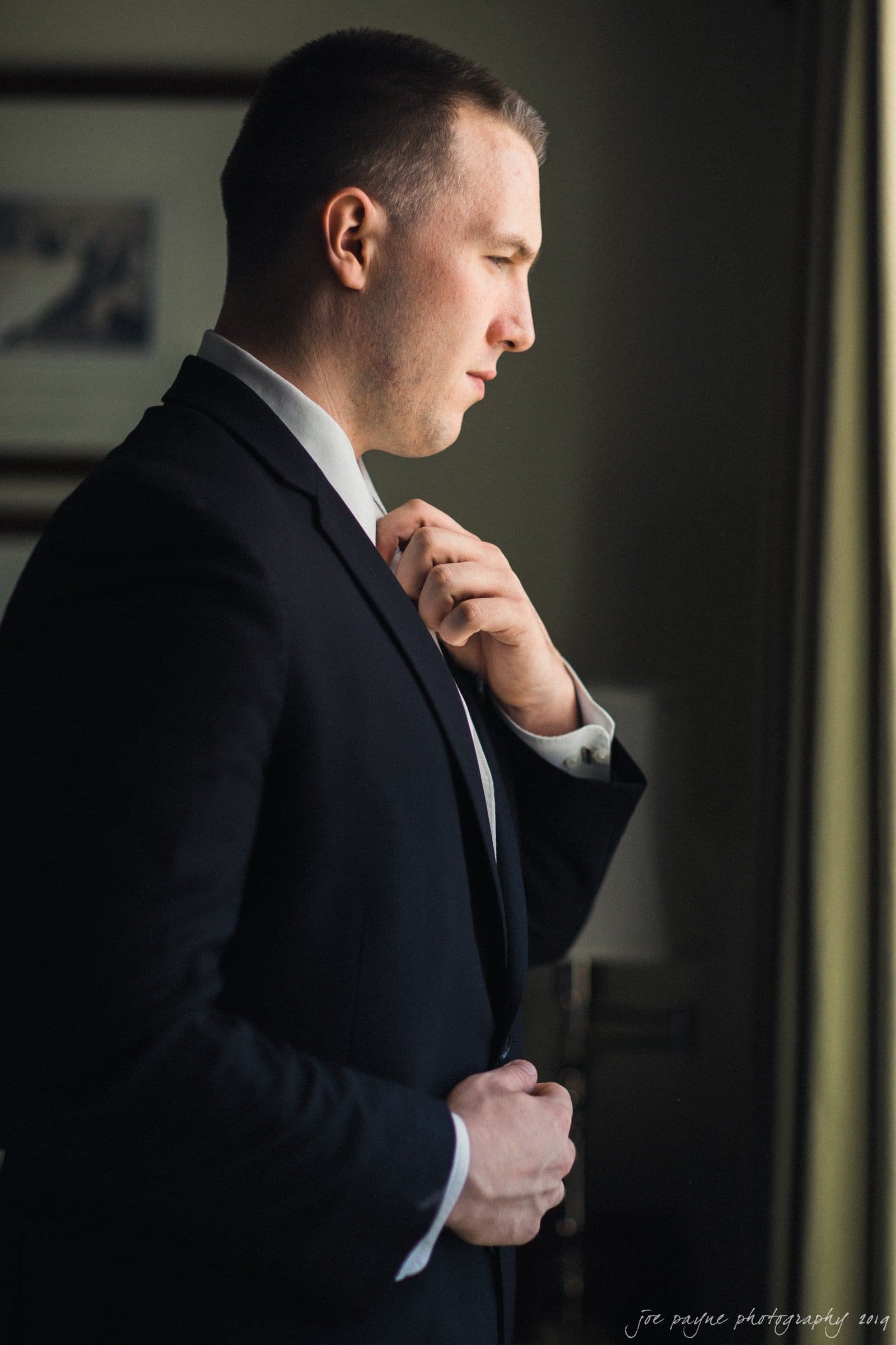 carolina inn & duke chapel wedding - kathleen & michael A man in a suit stands by a window, adjusting his tie. Soft light illuminates his profile, creating a contemplative mood. The rooms background is softly blurred, emphasizing his thoughtful expression.