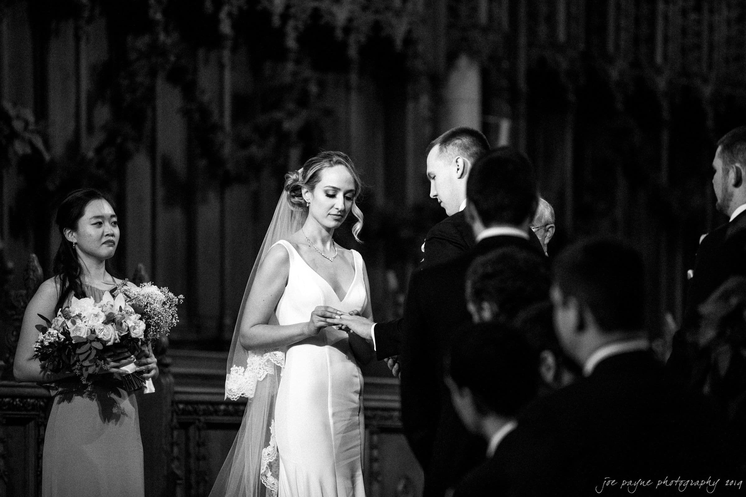 carolina inn & duke chapel wedding - kathleen & michael A black and white photo of a bride and groom exchanging rings during their wedding ceremony. The bride wears a long dress and veil, while the groom is in a dark suit. A bridesmaid holds a bouquet and watches the couple.