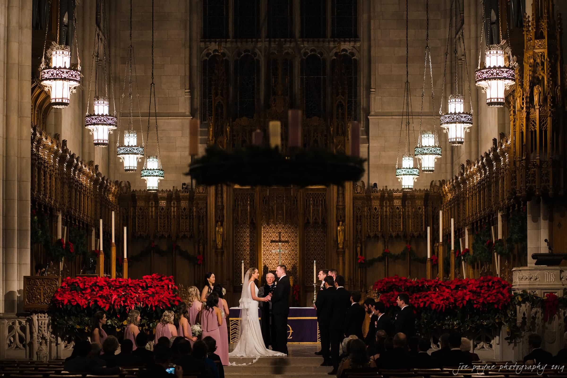 carolina inn & duke chapel wedding - kathleen & michael A couple exchanges vows at a wedding ceremony inside a grand cathedral. The scene is adorned with red poinsettias and lit by ornate chandeliers. Bridesmaids and groomsmen stand on either side, with a large audience observing.