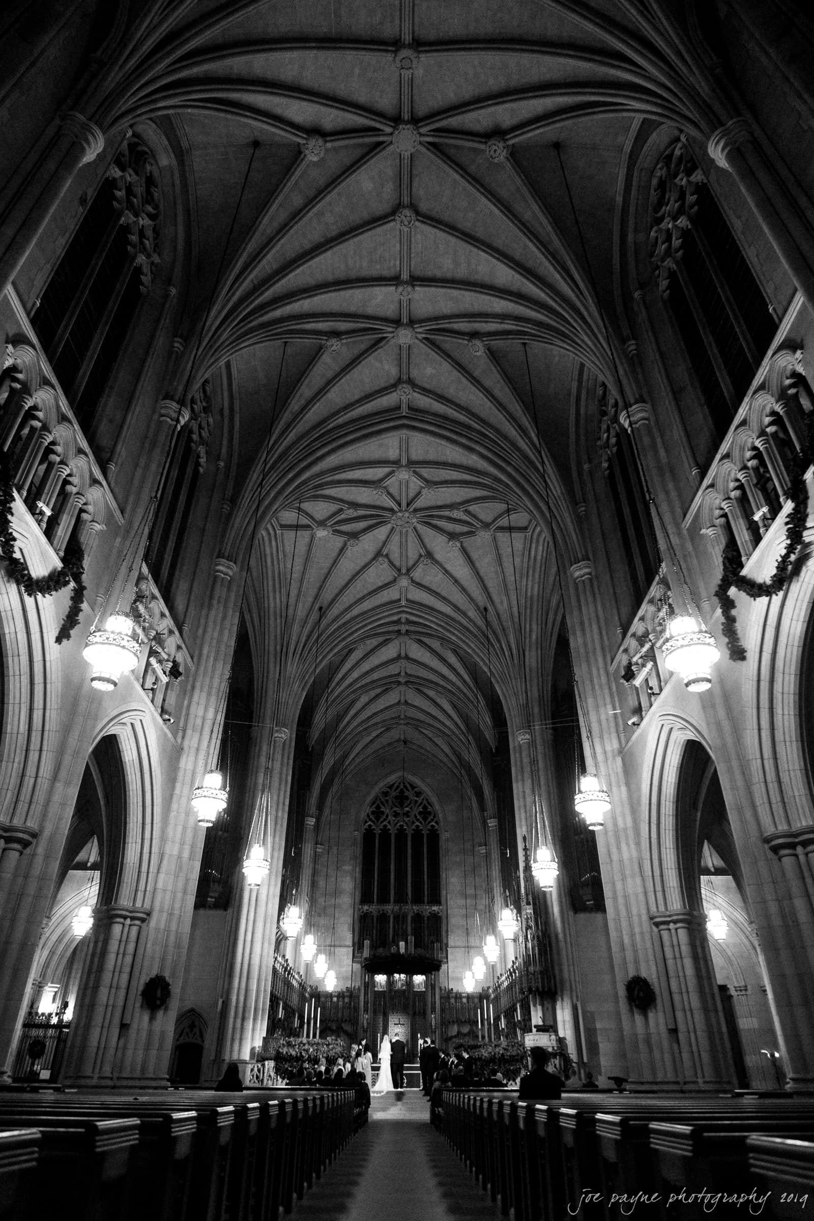 carolina inn & duke chapel wedding - kathleen & michael A black and white image of a grand, ornate church interior, featuring a vaulted ceiling and columns. A couple stands at the altar, surrounded by pews and illuminated by elegant hanging lights.