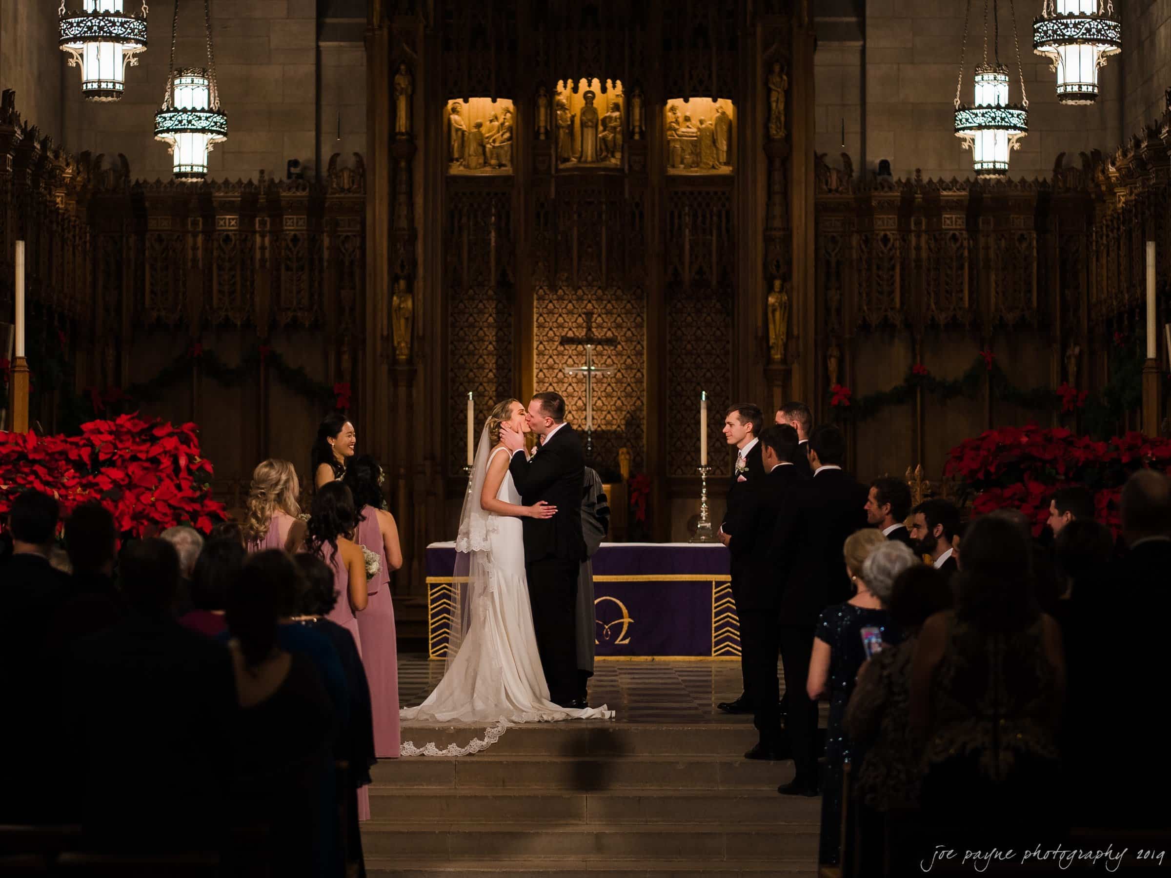 carolina inn & duke chapel wedding - kathleen & michael A bride and groom share a kiss at the altar in a dimly lit church. Bridesmaids in pink dresses and groomsmen in dark suits stand beside them. The altar is adorned with red poinsettias and candles, and an audience watches the ceremony.