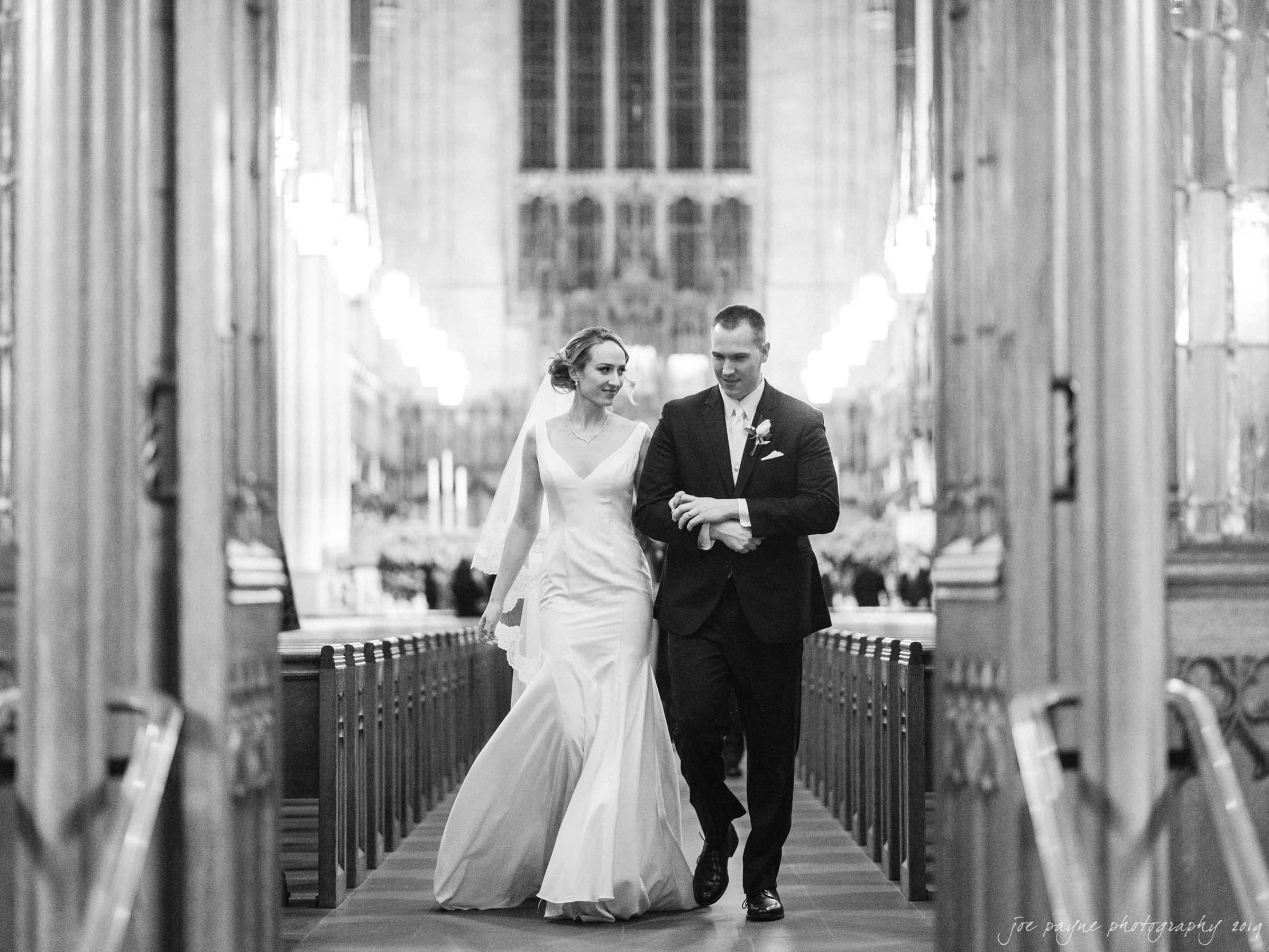 carolina inn & duke chapel wedding - kathleen & michael A bride and groom walk arm in arm down the aisle of an ornate, grand church lit with chandeliers. She wears a long, elegant gown and veil; he is in a dark suit. The setting is formal and romantic, capturing their joyous moment.