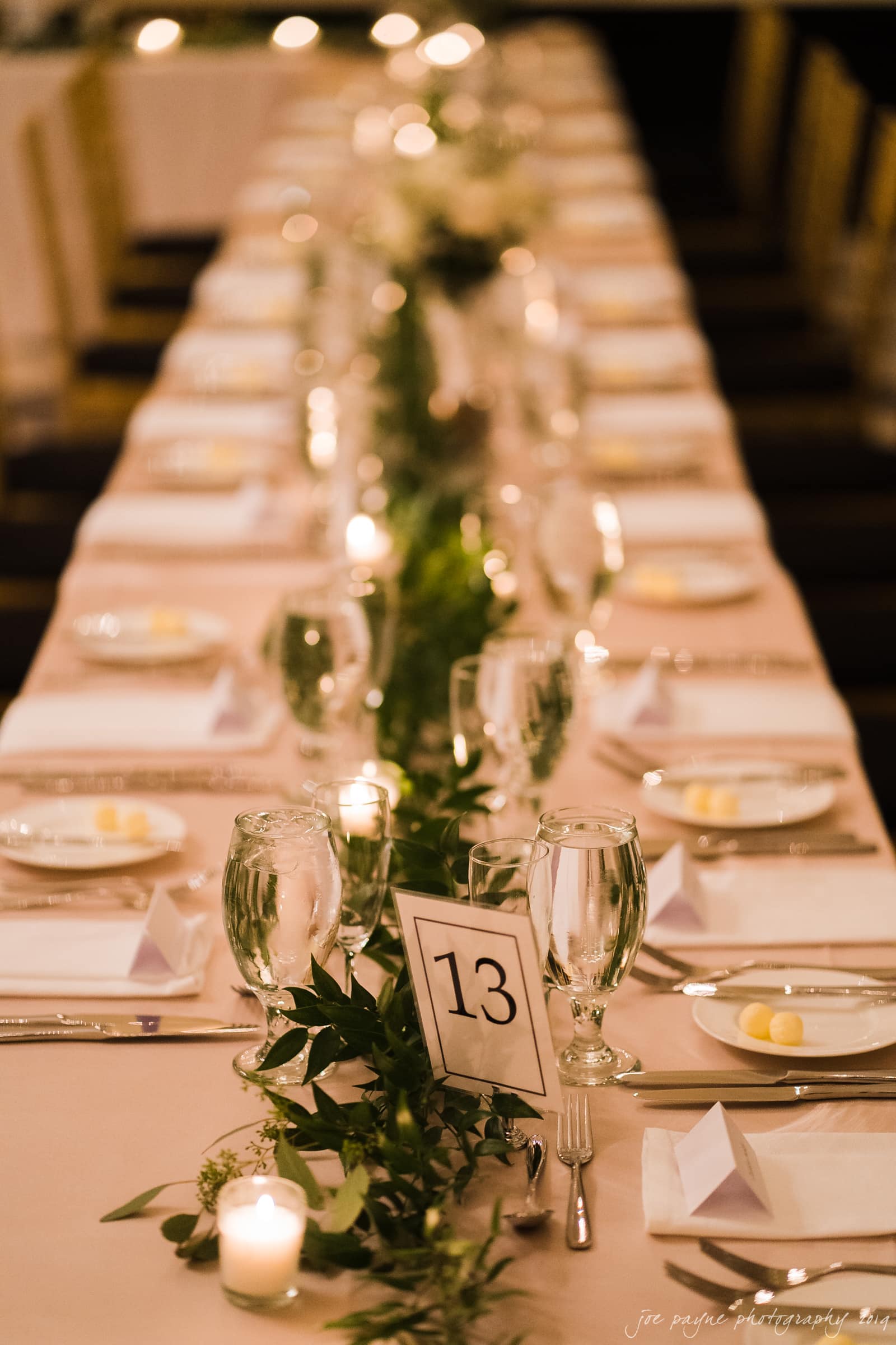 carolina inn & duke chapel wedding - kathleen & michael Elegant wedding reception table with a white tablecloth, set for guests. Decorated with green foliage, candles, and a table number 13. Each place has a white napkin, plate, and glassware, creating a sophisticated atmosphere.