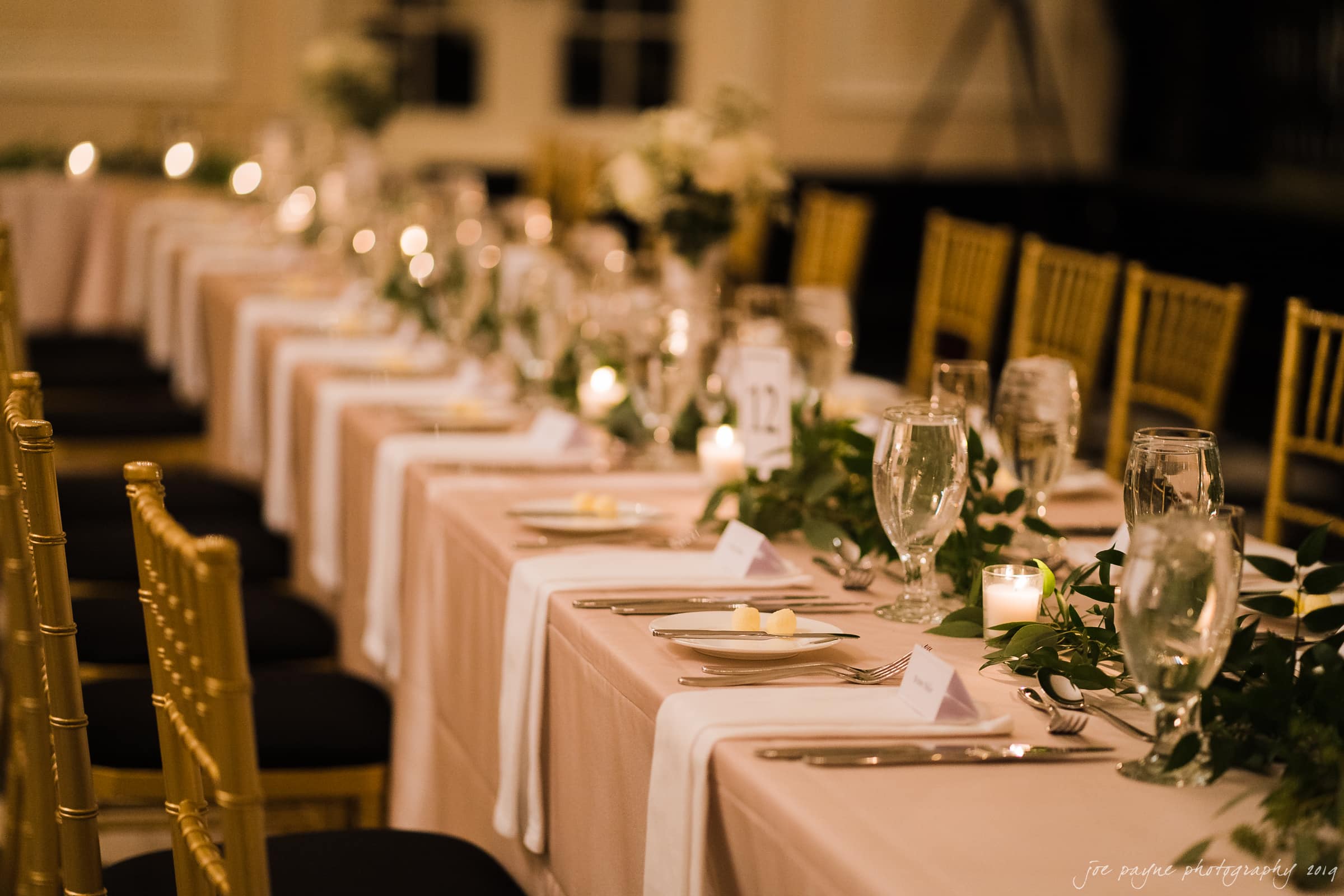 carolina inn & duke chapel wedding - kathleen & michael A long dining table is elegantly set with white plates, silverware, and wine glasses. A green garland and small candles decorate the center, creating a warm, intimate atmosphere. Gold chairs with black cushions are neatly aligned along the table.