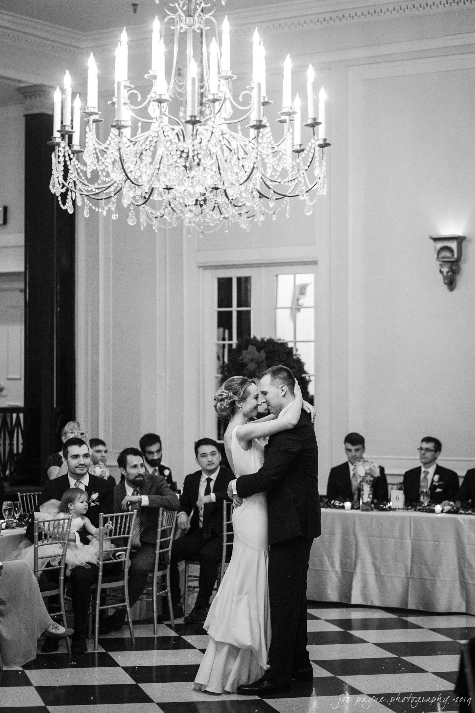 carolina inn & duke chapel wedding - kathleen & michael A couple shares a romantic dance under a grand chandelier in an elegant hall. Guests sitting at tables watch the scene, surrounded by sophisticated decor and a large floral arrangement. The mood is joyful and intimate.