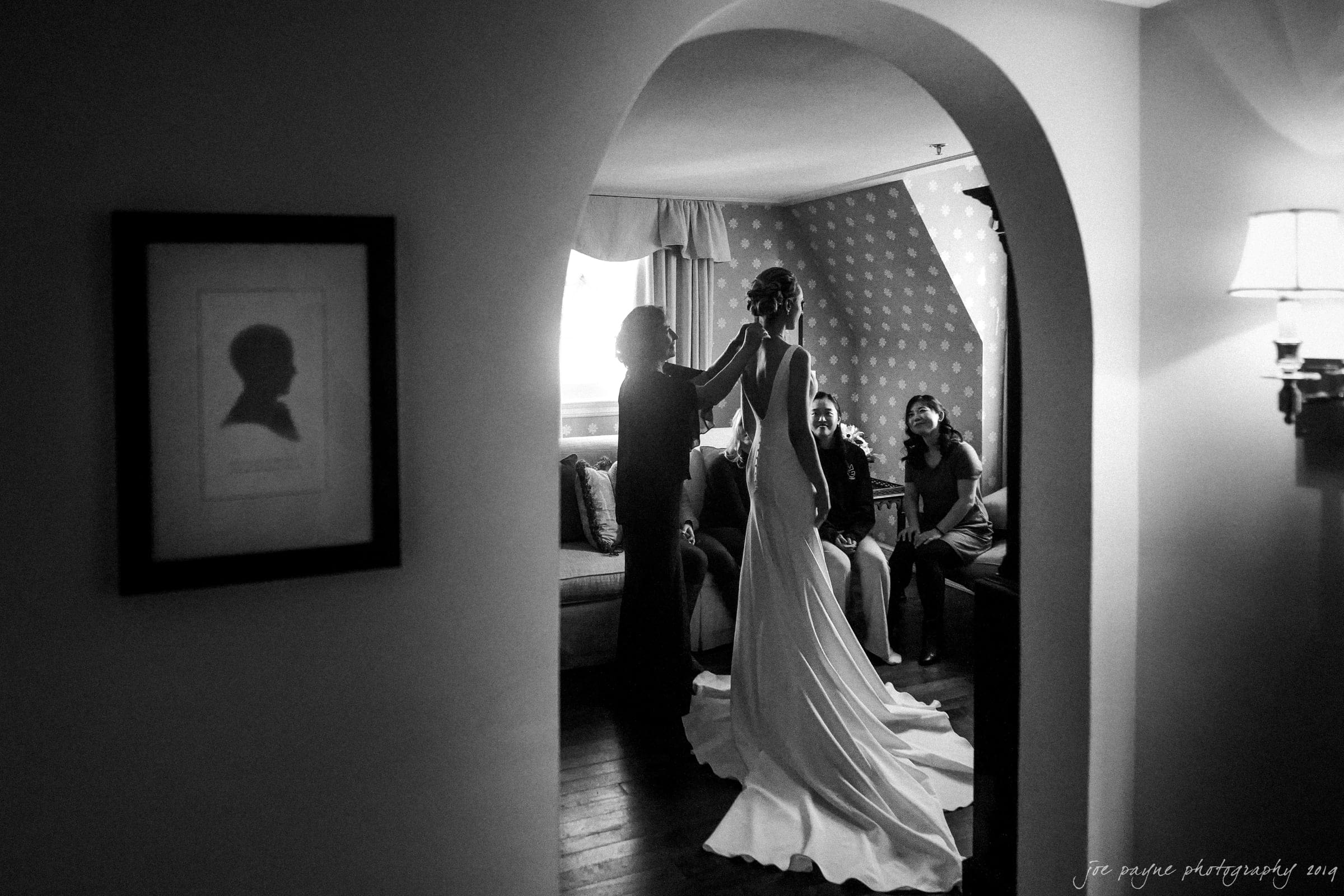 carolina inn & duke chapel wedding - kathleen & michael A silhouetted bride in an elegant gown stands gracefully in a dimly lit room, as two seated women and an older woman admire her. The scene is framed by an arched doorway, creating an intimate and timeless atmosphere.