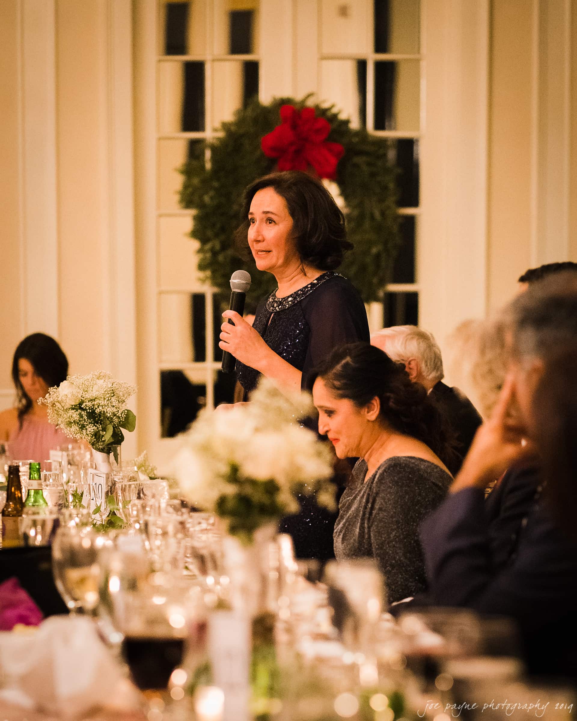 carolina inn & duke chapel wedding - kathleen & michael A woman stands and speaks into a microphone at a formal event. She is surrounded by seated guests at a table adorned with flowers and glasses. A large wreath with a red bow decorates the wall behind her.