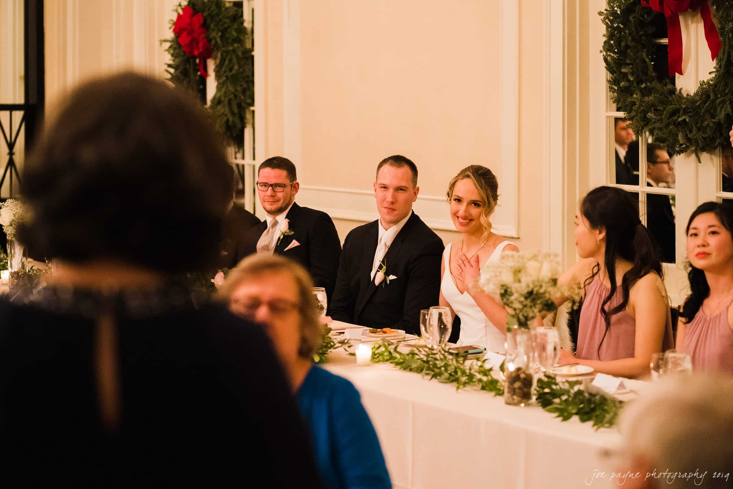 carolina inn & duke chapel wedding - kathleen & michael A wedding reception scene with a bride and groom seated at a table, adorned with flowers and candles. They are smiling and surrounded by friends. A guest speaks to them, while holiday wreaths decorate the walls.