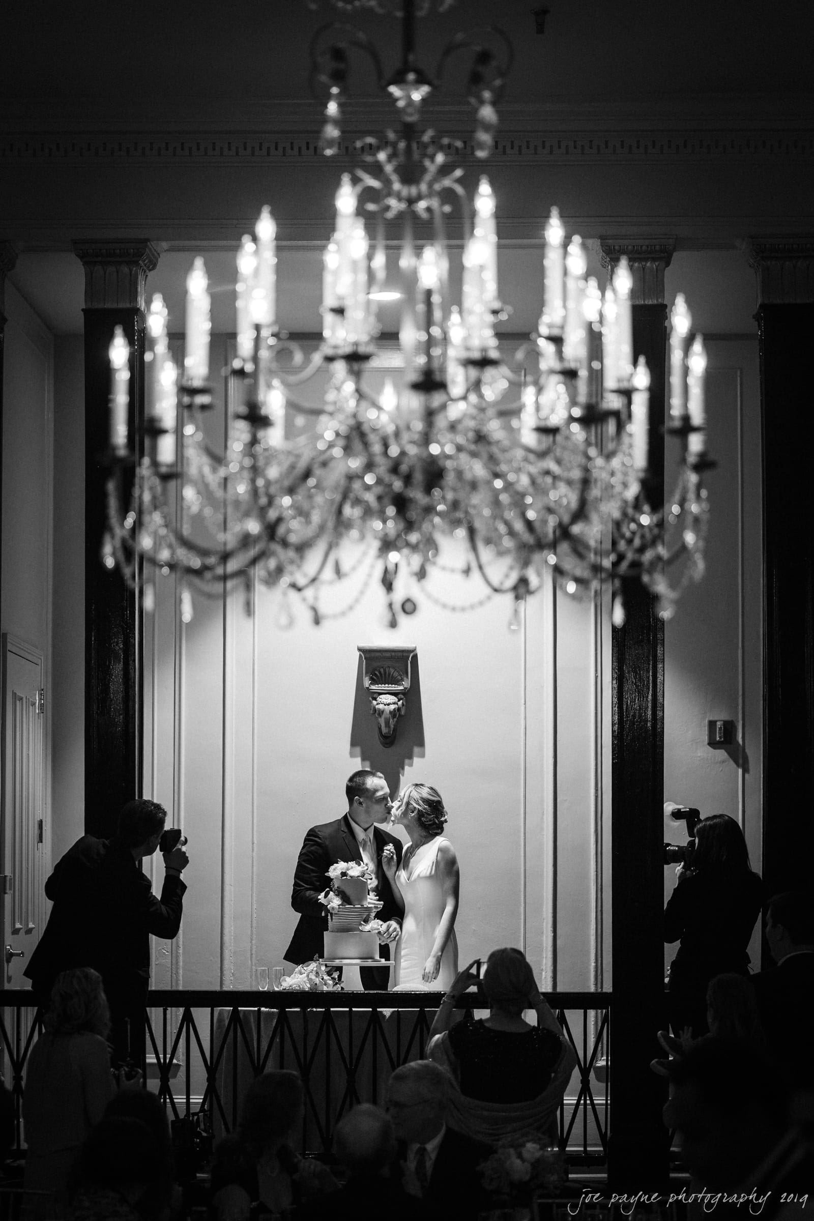 carolina inn & duke chapel wedding - kathleen & michael A bride and groom stand behind a wedding cake in a dimly lit room. A large chandelier dominates the foreground. Photographers capture the moment as guests look on. The setting is elegant and intimate.