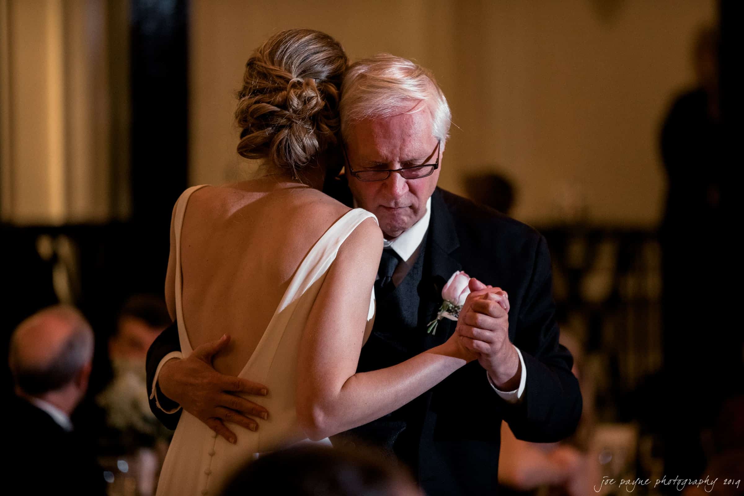 carolina inn & duke chapel wedding - kathleen & michael A woman in a white dress dances closely with an older man in a suit, who holds a pink rose boutonniere. The setting appears to be a formal indoor event. The mood is intimate and emotional.