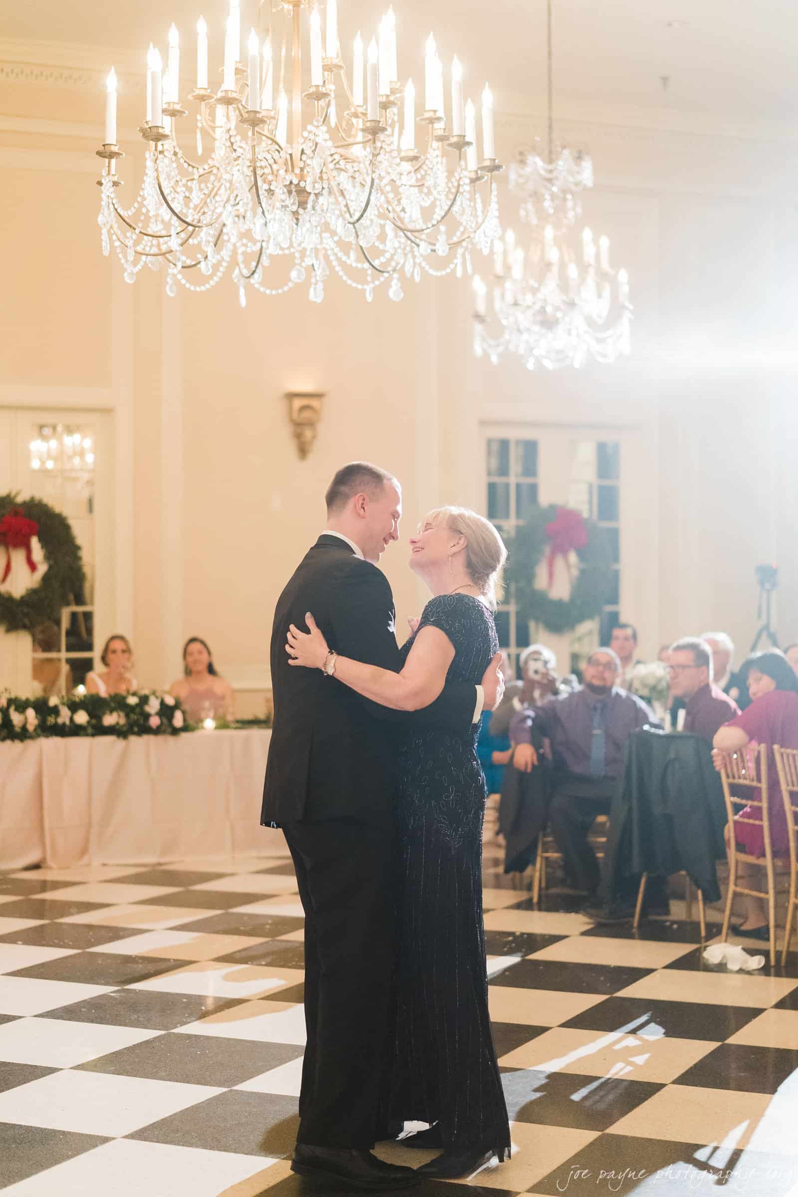 carolina inn & duke chapel wedding - kathleen & michael A couple dances closely on a black and white checkered floor under a large chandelier. In the background, guests seated at decorated tables watch the dance, with a wreath hanging on a wall.