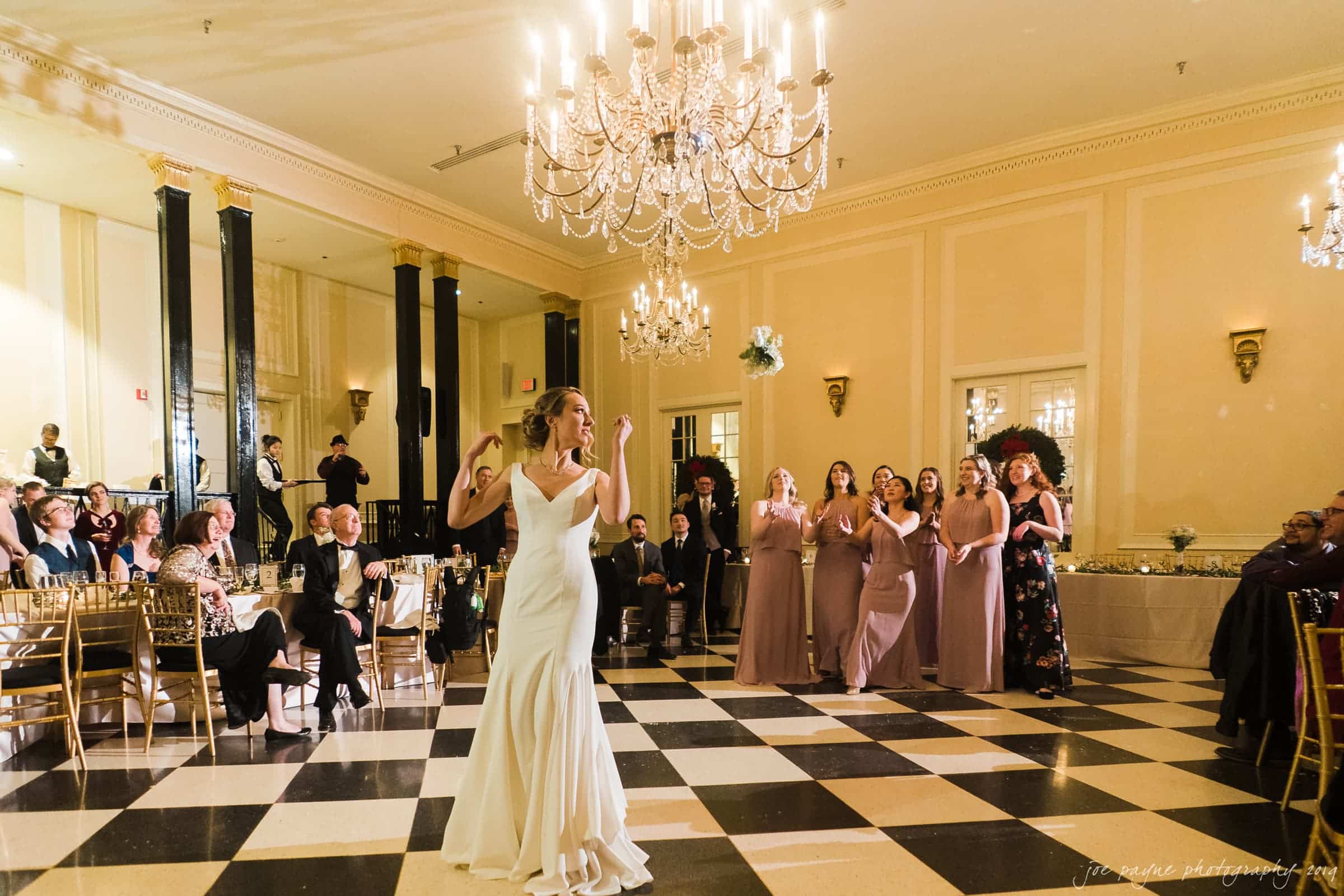 carolina inn & duke chapel wedding - kathleen & michael A bride in a white gown dances under a chandelier on a checkered floor, surrounded by seated guests. Bridesmaids in mauve dresses watch happily. The room is elegantly decorated with chandeliers and tall mirrors.