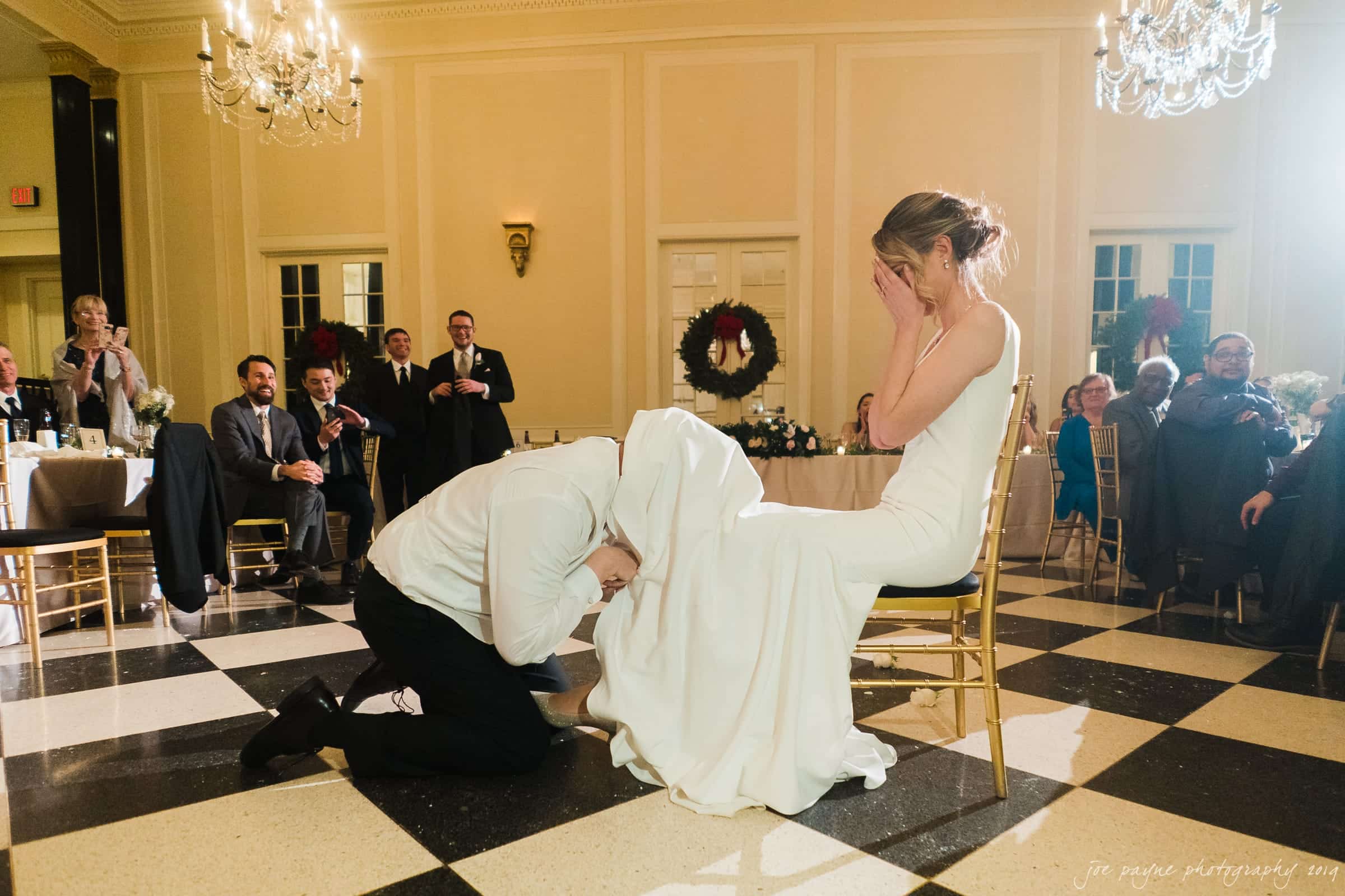 carolina inn & duke chapel wedding - kathleen & michael A bride and groom participate in a wedding tradition. The groom kneels in front of the seated bride, playfully reaching under her dress. Guests watch and smile in a warmly lit banquet hall with checkered floors and chandeliers.