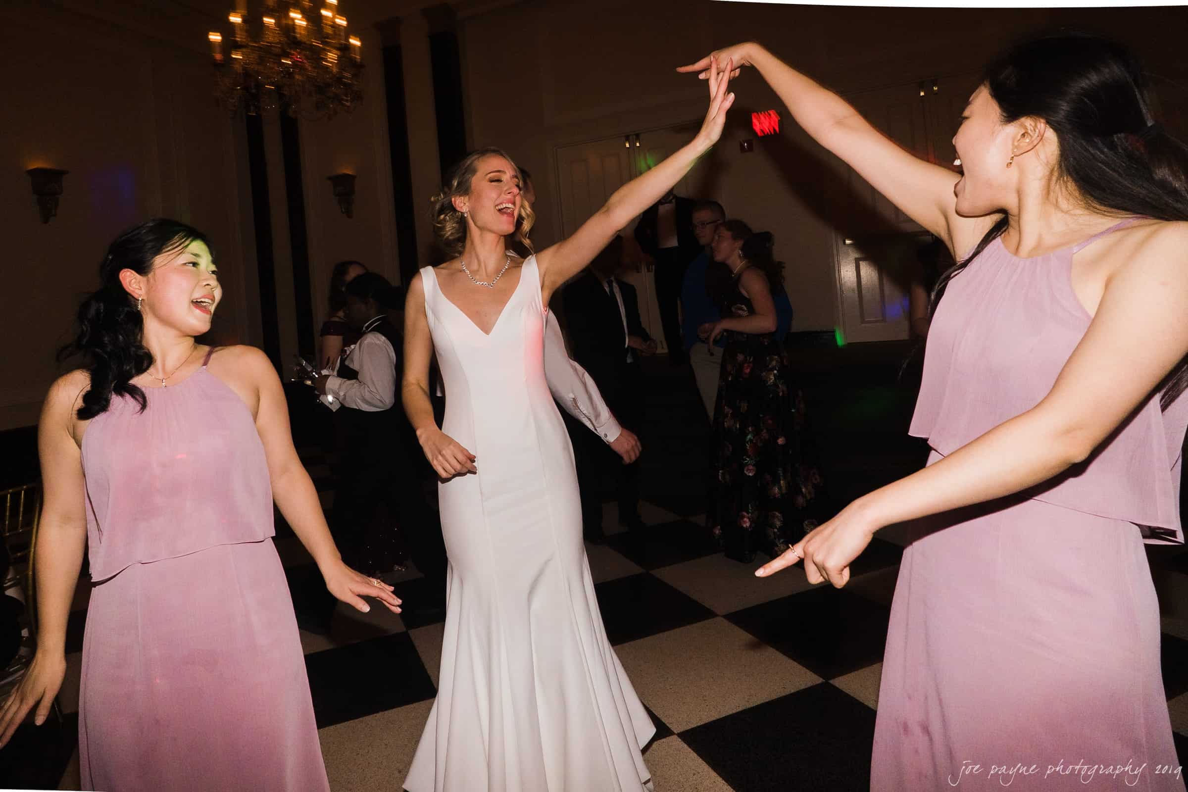 carolina inn & duke chapel wedding - kathleen & michael Three women happily dancing at an indoor event. The woman in the center wears a white dress, while the other two wear matching purple dresses. The room has a checkered floor and a chandelier, with several people in the background.