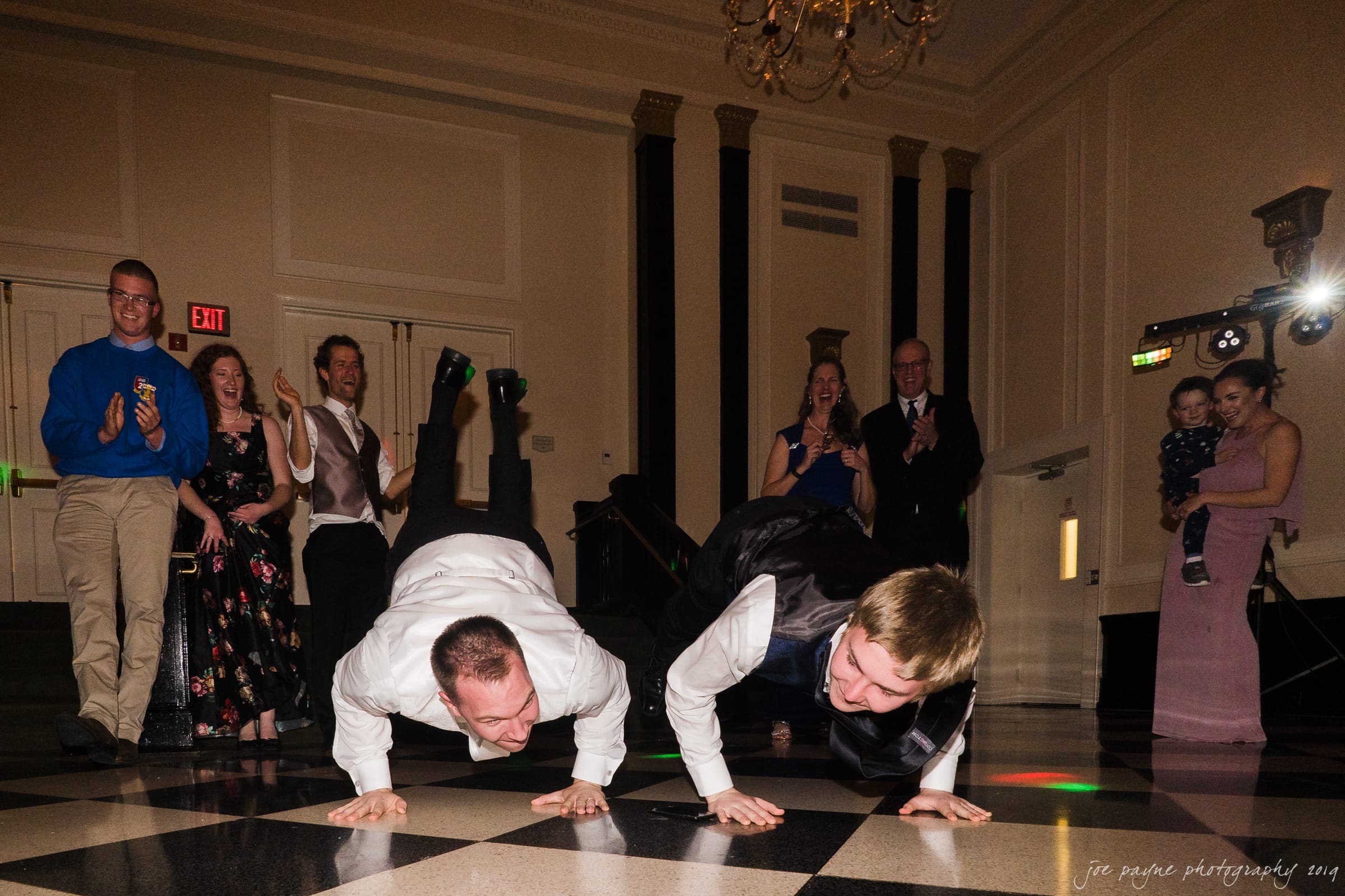 carolina inn & duke chapel wedding - kathleen & michael Two men, wearing dress shirts and vests, are doing push-ups on the floor at a lively indoor event. A group of six people stands in the background, smiling and clapping, with some holding drinks. The room has elegant decor and dim lighting.