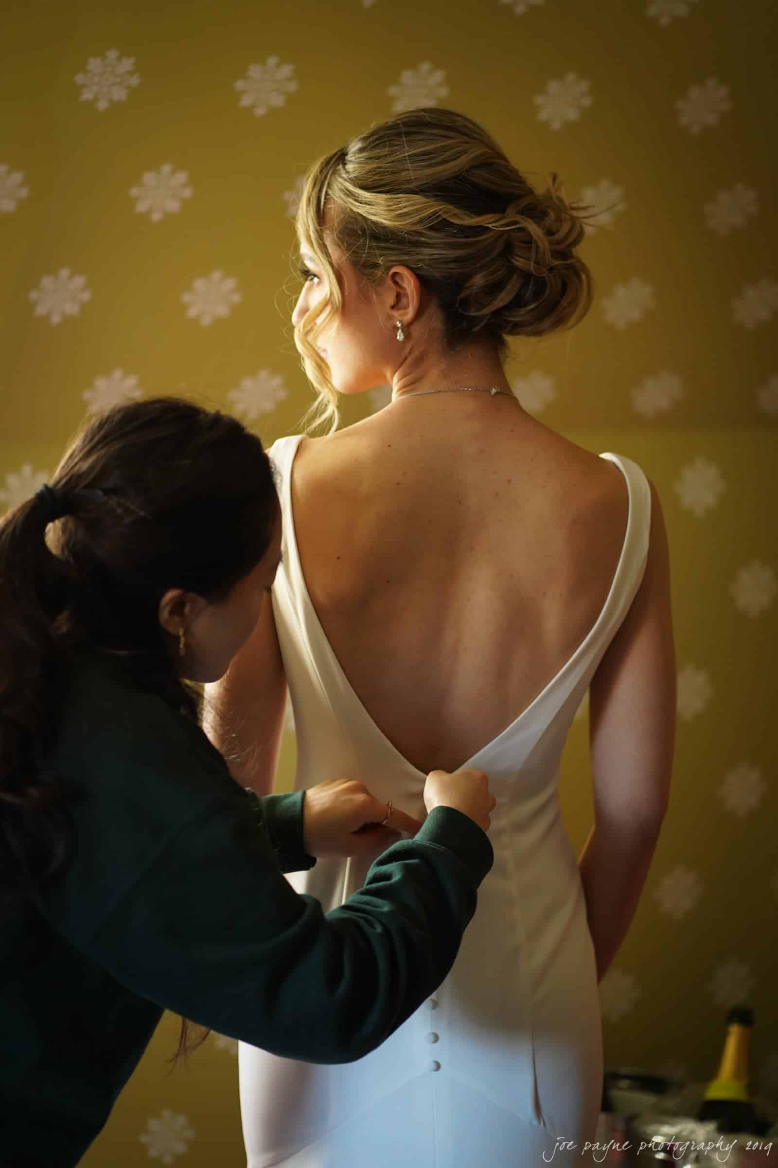 carolina inn & duke chapel wedding - kathleen & michael A bride in a sleeveless white gown is getting assistance from another person with dark hair, who is adjusting the dresss low back. The scene is set against a green floral-patterned wall, with soft lighting highlighting the brides hairstyle.