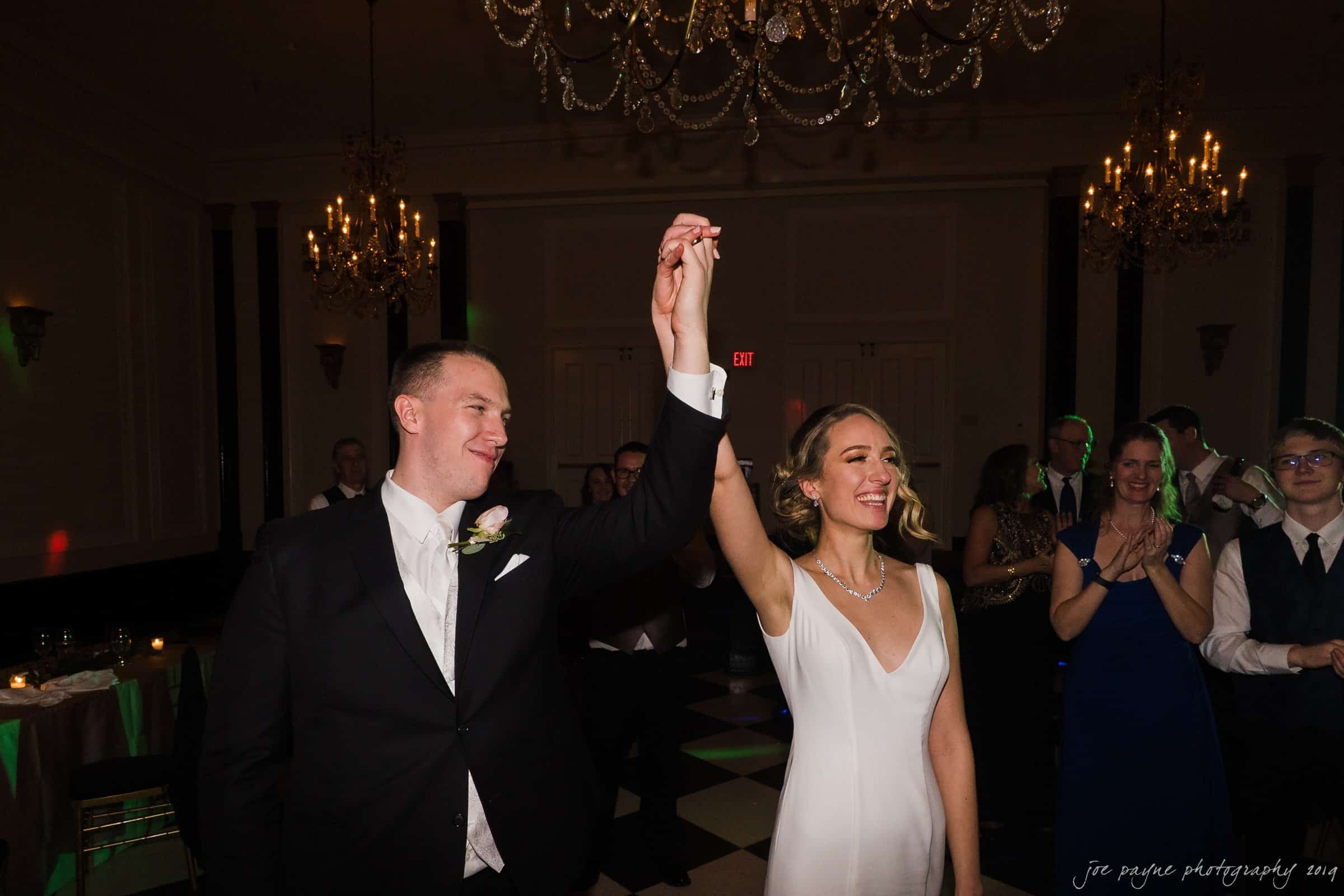 carolina inn & duke chapel wedding - kathleen & michael A couple in formal attire, holding hands and smiling, walks through a ballroom under elegant chandeliers. Guests in the background clap and smile, celebrating the moment.