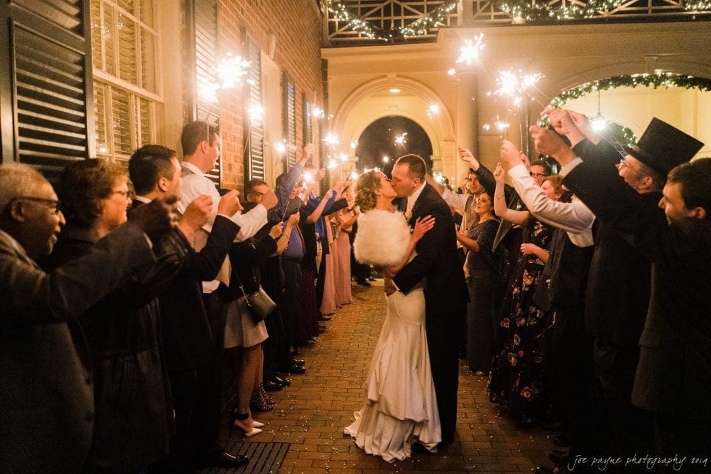A bride and groom share a kiss under an archway of sparklers, held by wedding guests forming two lines. The scene takes place in a warmly lit corridor with brick flooring and decorative lights above.
