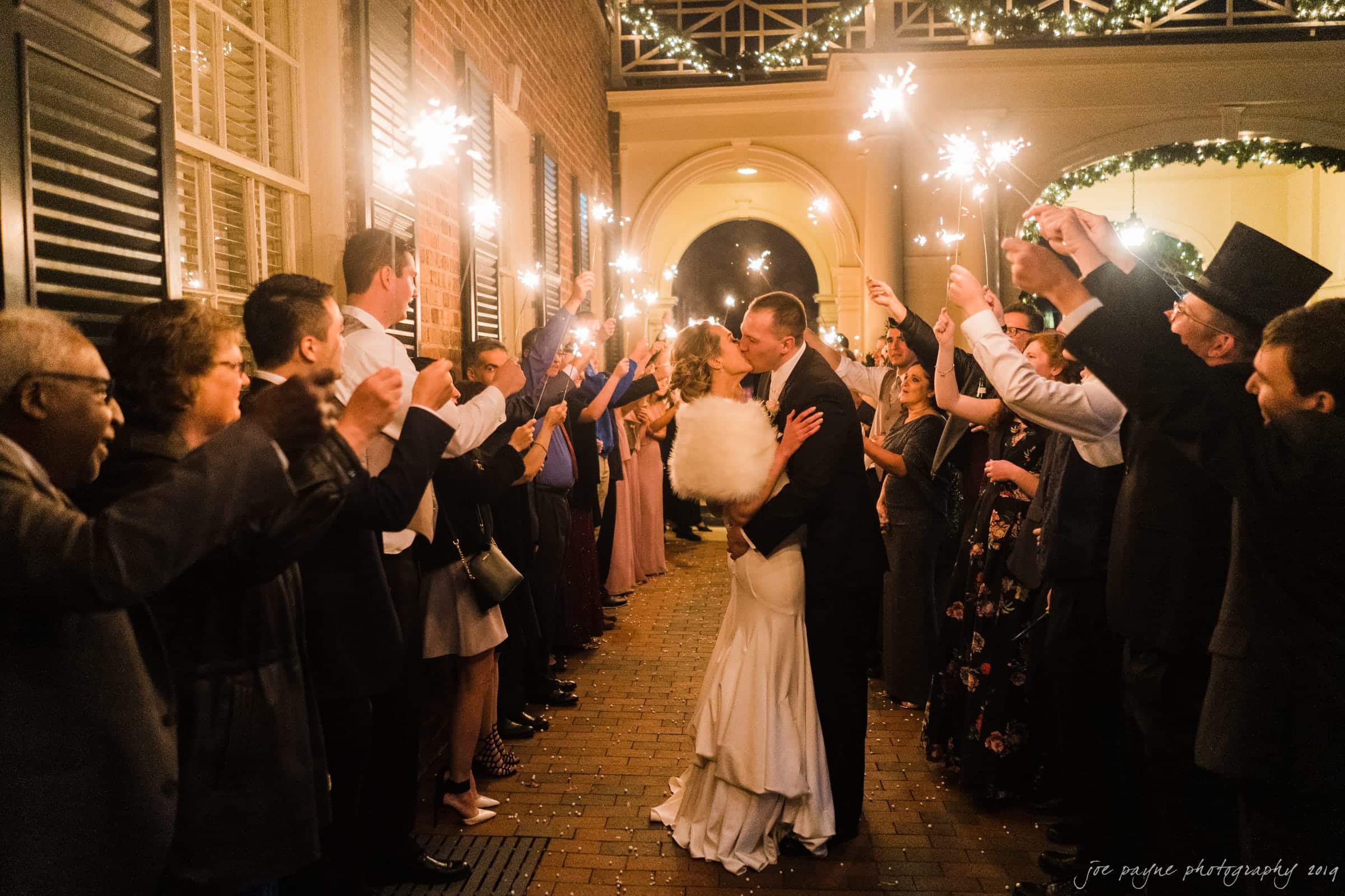 carolina inn & duke chapel wedding - kathleen & michael A bride and groom share a kiss under an archway of sparklers, held by wedding guests forming two lines. The scene takes place in a warmly lit corridor with brick flooring and decorative lights above.
