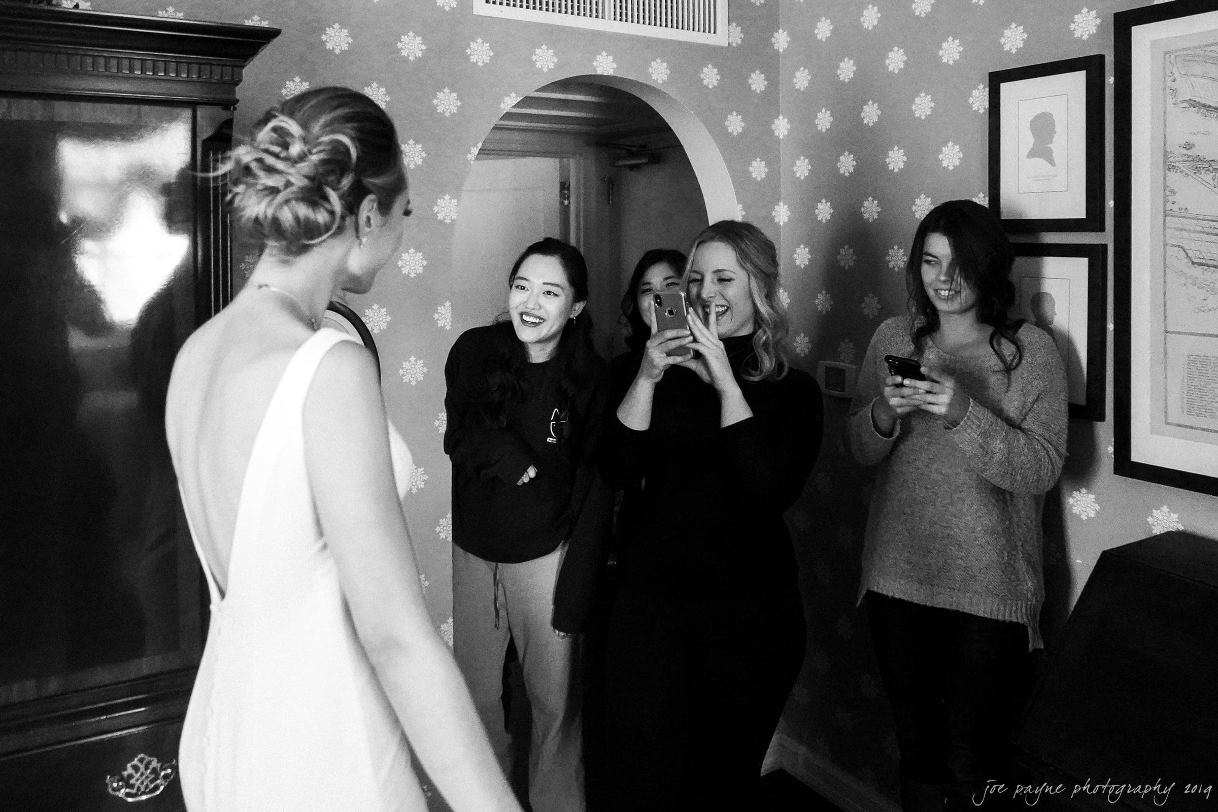 carolina inn & duke chapel wedding - kathleen & michael A bride in a wedding dress stands in a decorated room, smiling at four women. Two women are taking photos with their phones, one woman is laughing, and another is smiling warmly at the bride. The setting looks cheerful and intimate.