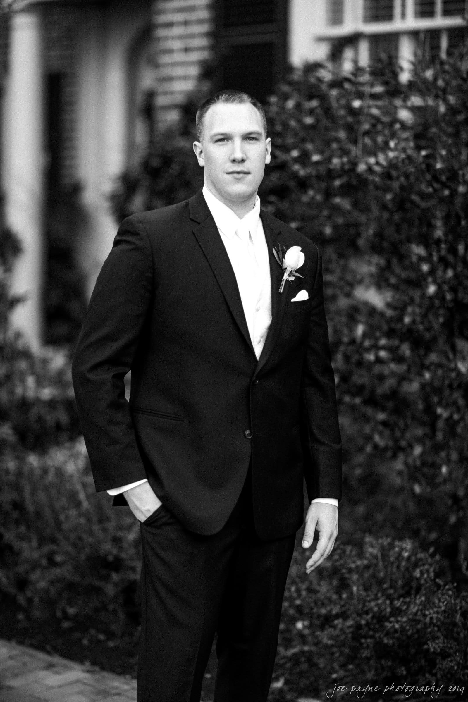 carolina inn & duke chapel wedding - kathleen & michael A groom in a suit with a boutonniere stands outdoors, posing for a portrait. The black and white image features a background of greenery and a building.