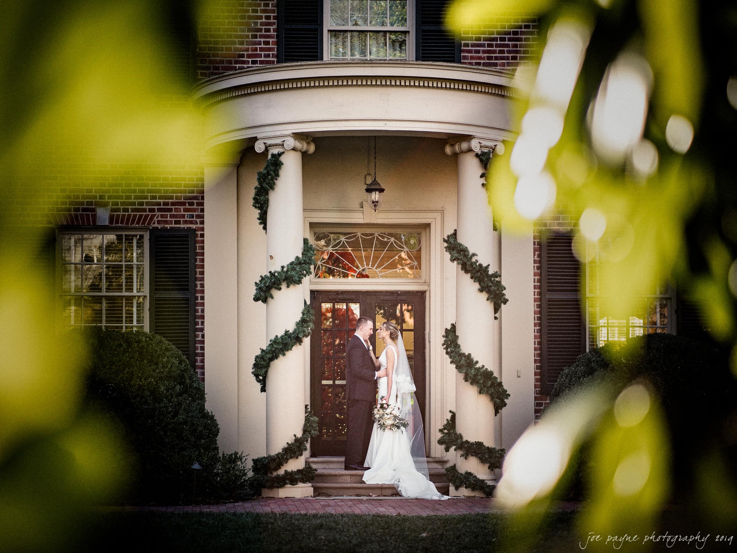 carolina inn & duke chapel wedding - kathleen & michael A bride and groom share a kiss in front of a grand entrance, decorated with garlands, on a brick building. The scene is framed by blurred green leaves, with warm lighting enhancing the romantic atmosphere.
