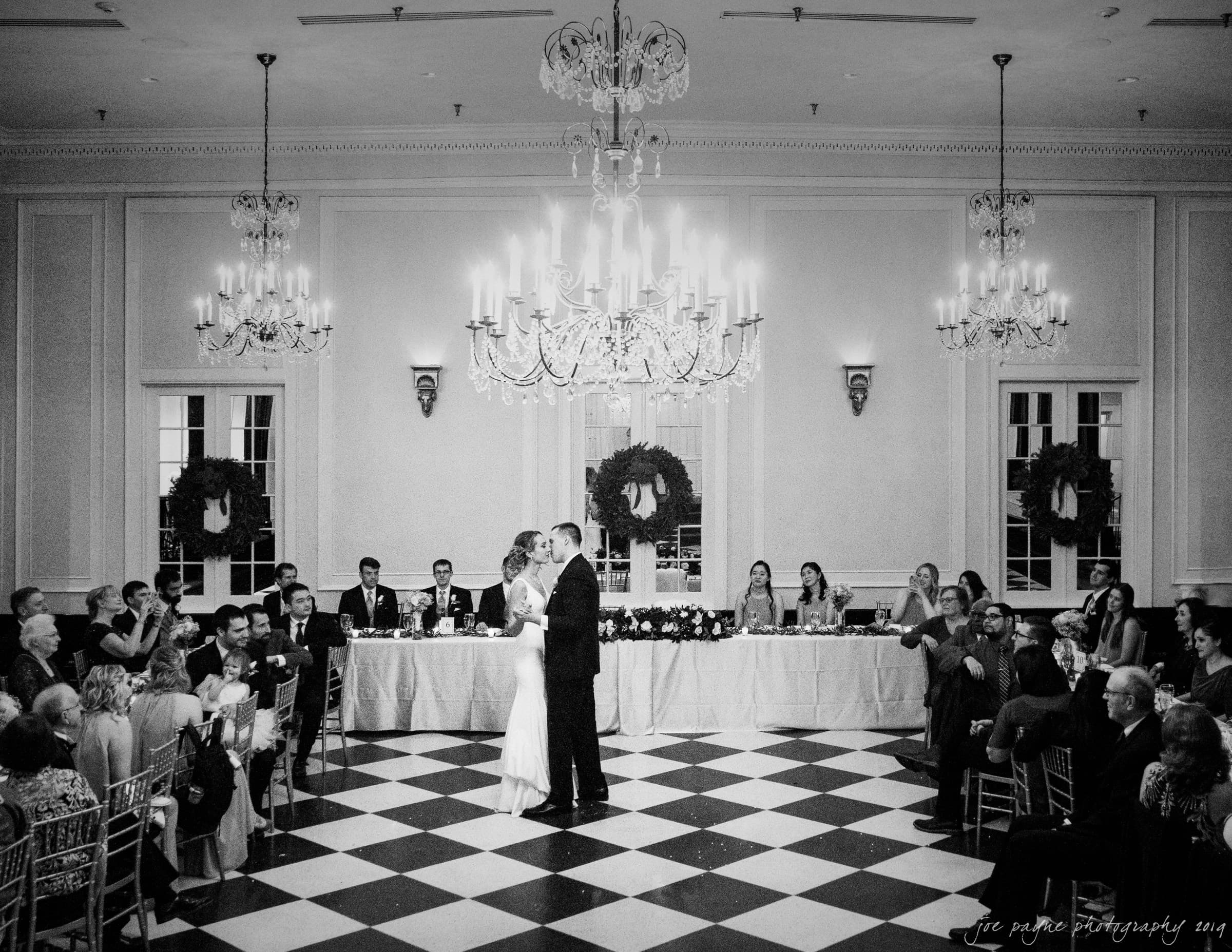 carolina inn & duke chapel wedding - kathleen & michael A couple shares their first dance in a grand ballroom. The room features chandeliers, wreaths, and a checkered floor. Guests are seated at tables and a long banquet table is visible in the background. The black-and-white photo captures an elegant ambiance.