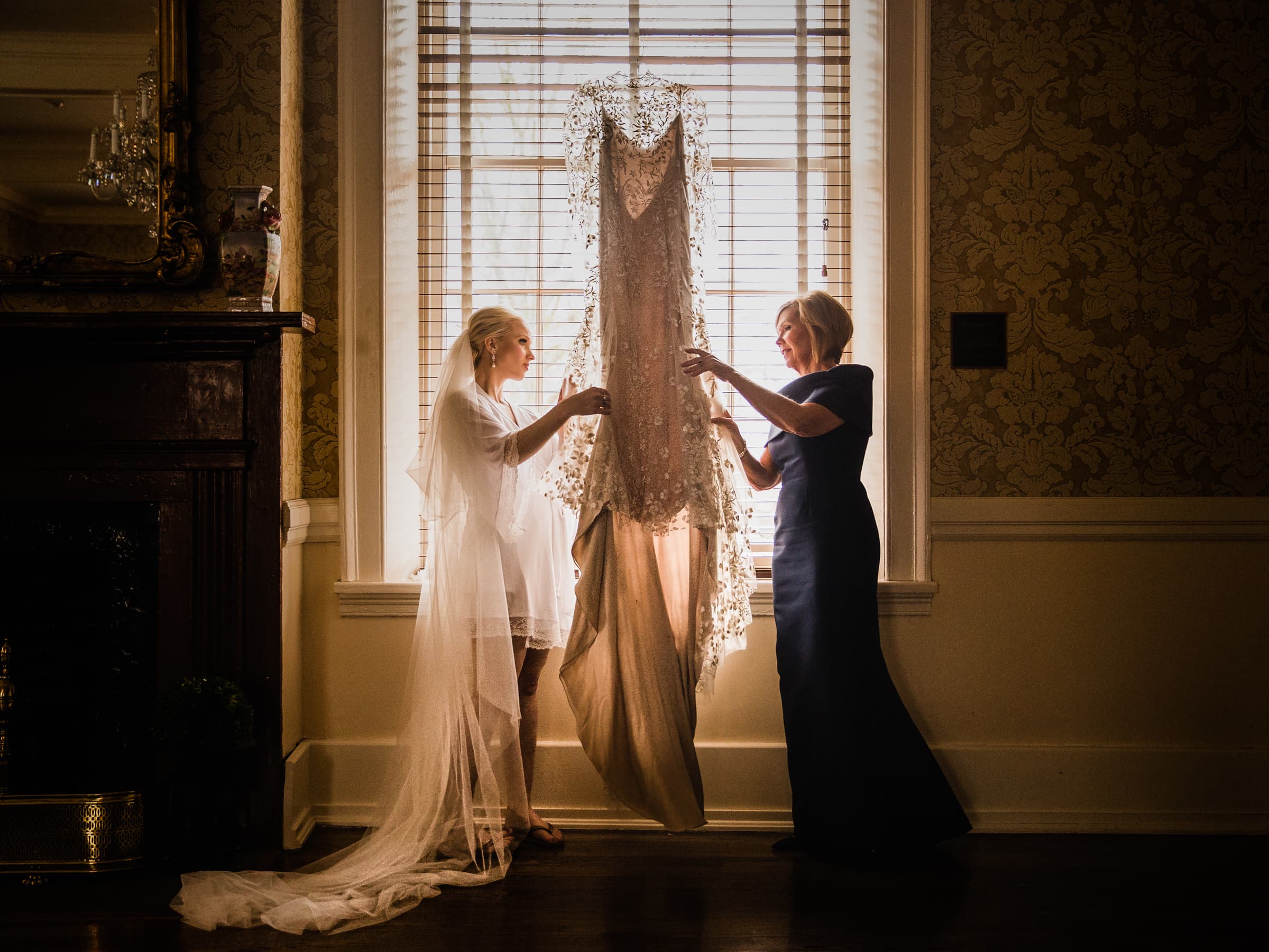 Two women stand by a window, engrossed in wedding photography planning. One wears a white robe and veil, the other a navy blue dress. Sunlight filters through the blinds, casting a glow on the wedding dress hanging elegantly from the curtain rod.