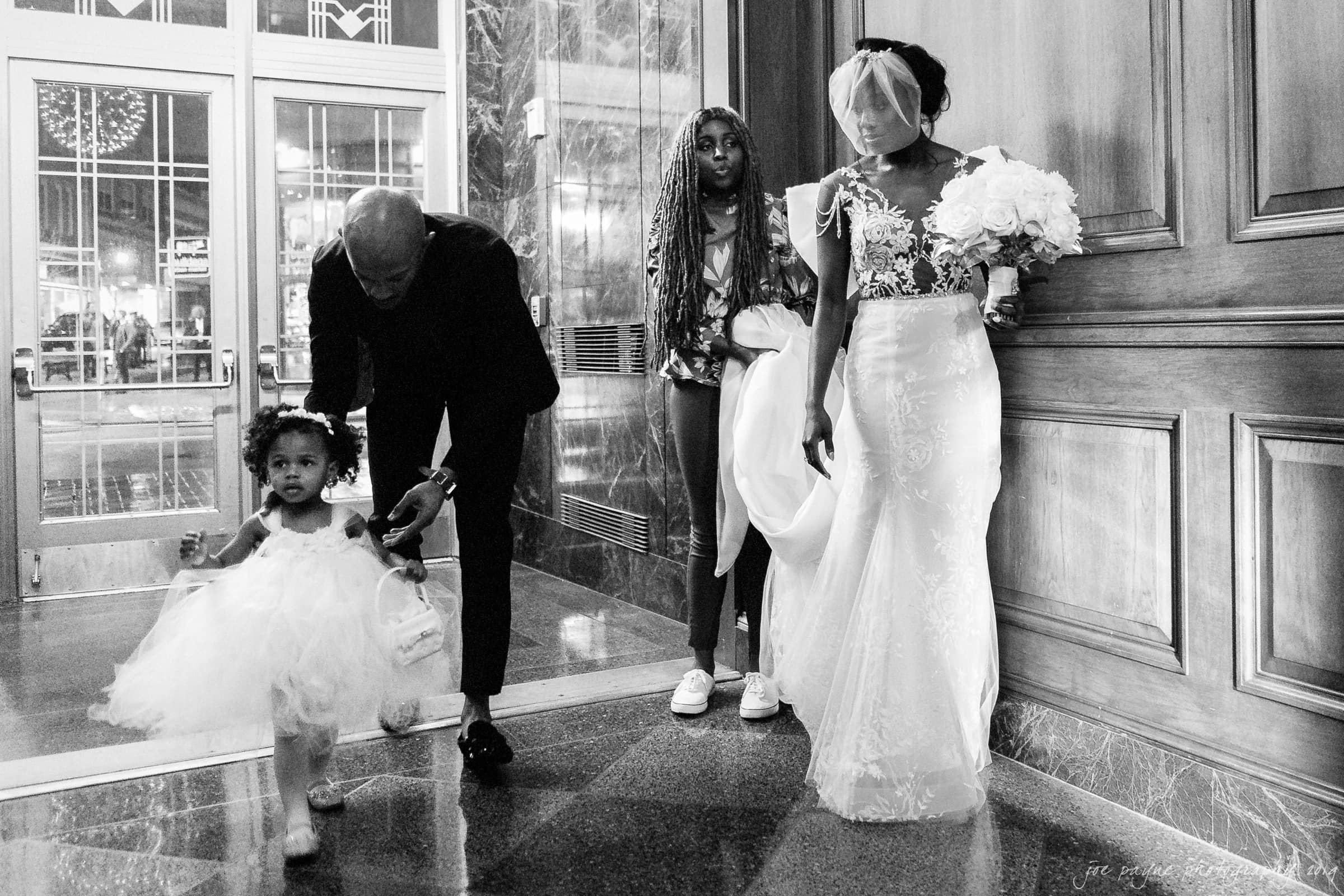 21c museum hotel durham wedding - kortne & terry A black and white photo of a bride holding a bouquet, a young girl in a white dress walking ahead, and a man helping her. Another young person stands nearby. They are in a hallway with reflections on the floor.