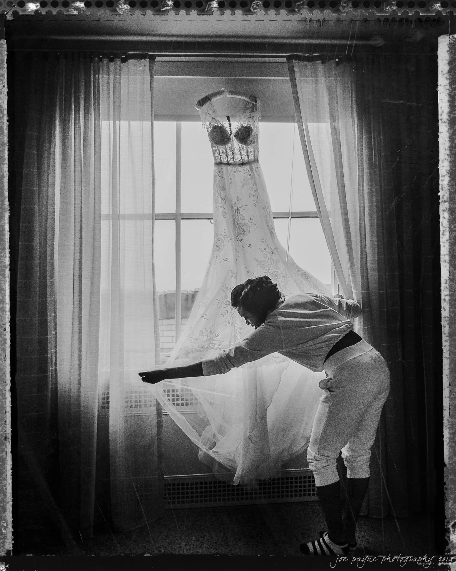 21c museum hotel durham wedding - kortne & terry A person adjusts the curtain near a window where a wedding dress is hanging. The dress has intricate details and the silhouette of the person adds to the composition. The scene is in black and white, creating a classic, timeless feel.
