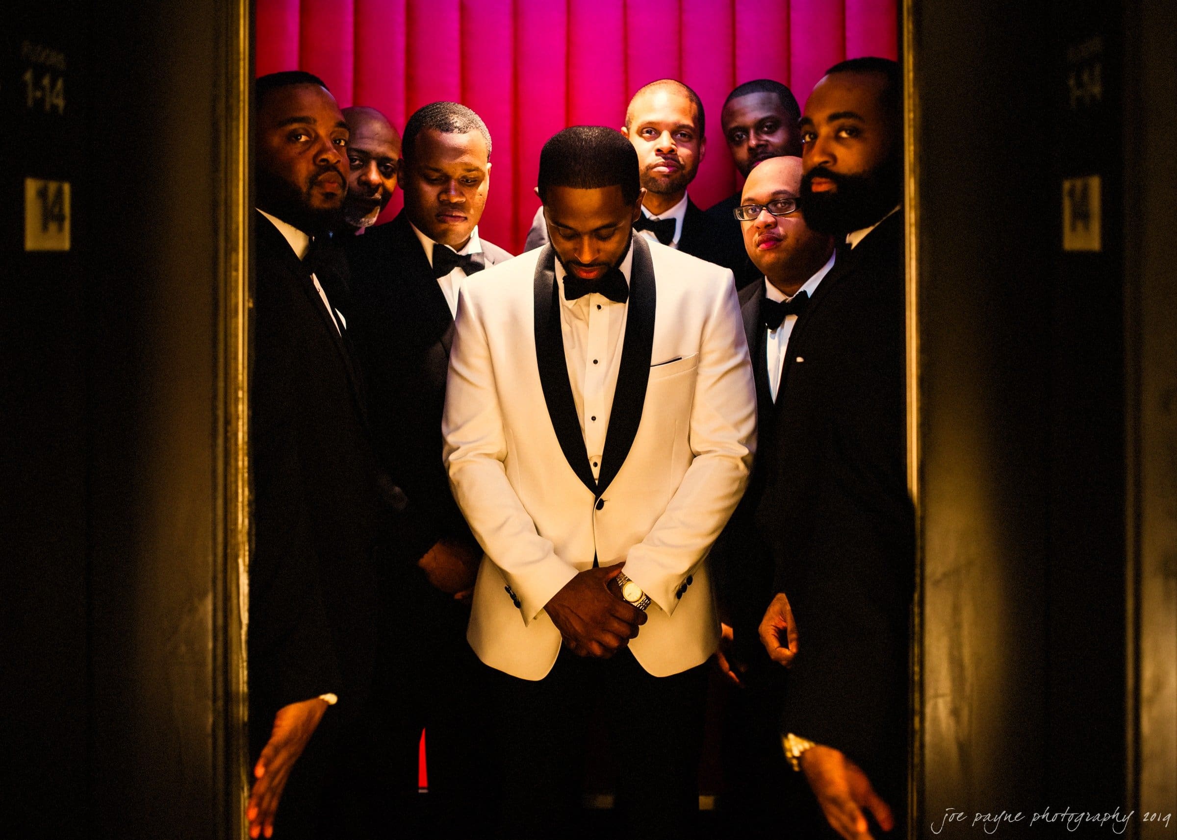 21c museum hotel durham wedding - kortne & terry A group of eight men dressed in tuxedos stand in an elevator with red walls. The man in the center wears a white jacket and looks down, while the others wear black jackets and look forward.
