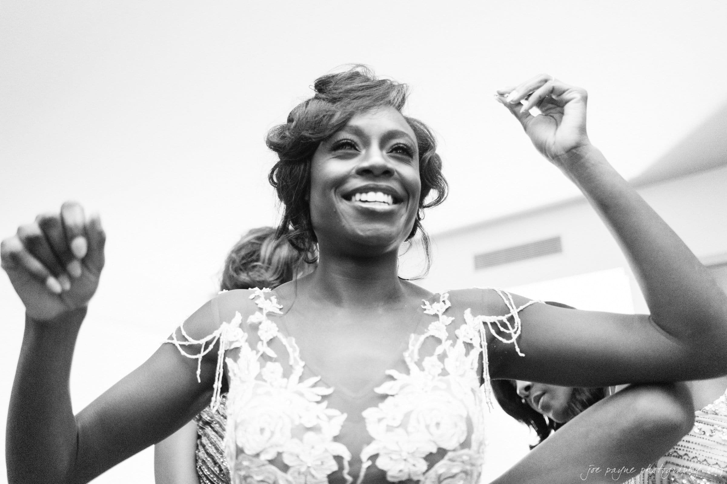 21c museum hotel durham wedding - kortne & terry A joyful bride in a lace wedding dress celebrates, raising her arms with a beaming smile. The black and white image captures her in a moment of happiness, surrounded by a soft, blurred background.