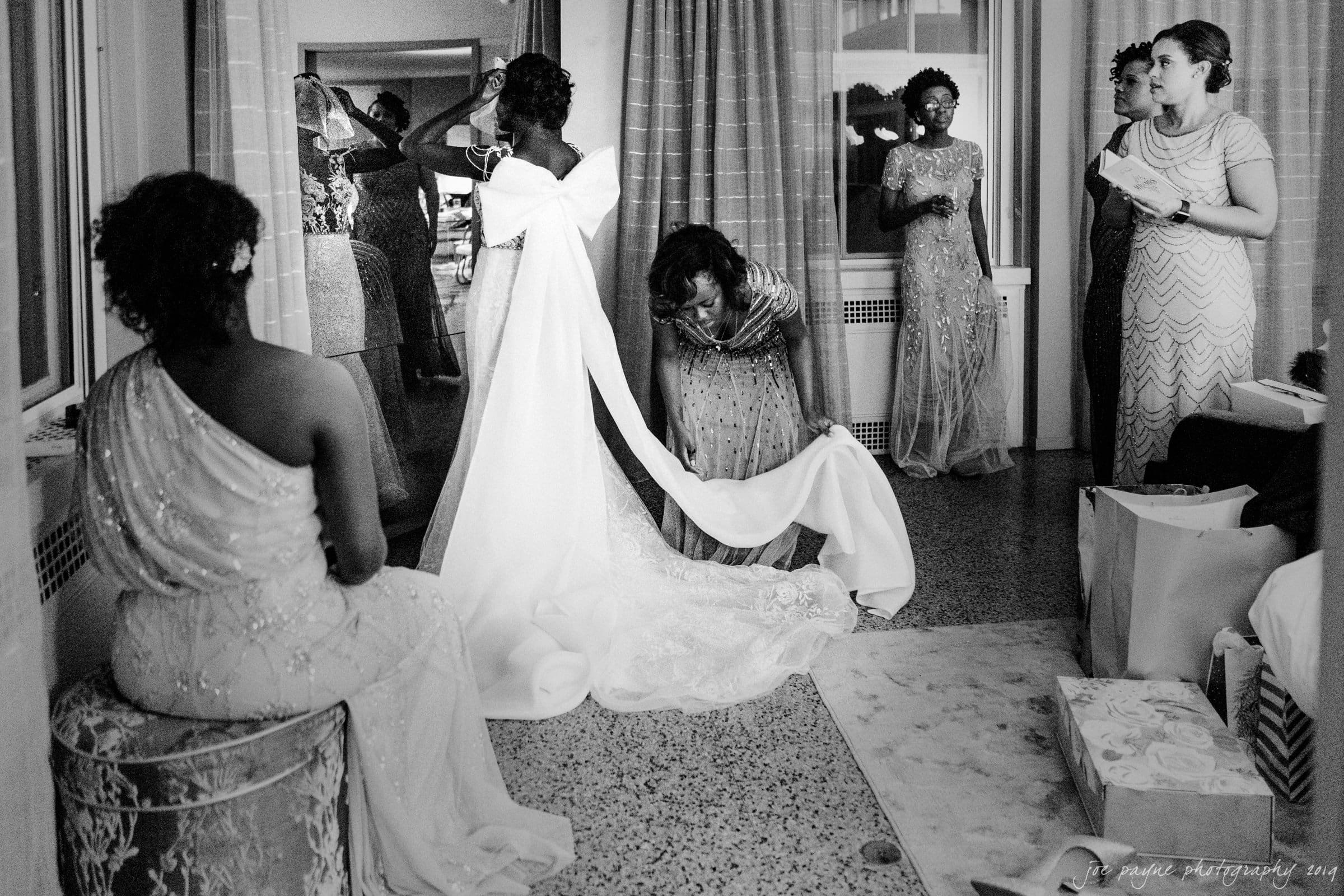 21c museum hotel durham wedding - kortne & terry A bride adjusts her off-the-shoulder gown while surrounded by women in elegant dresses. One woman helps arrange the train of the dress. The room has large windows and gift boxes on the floor, creating a lively and joyful atmosphere.