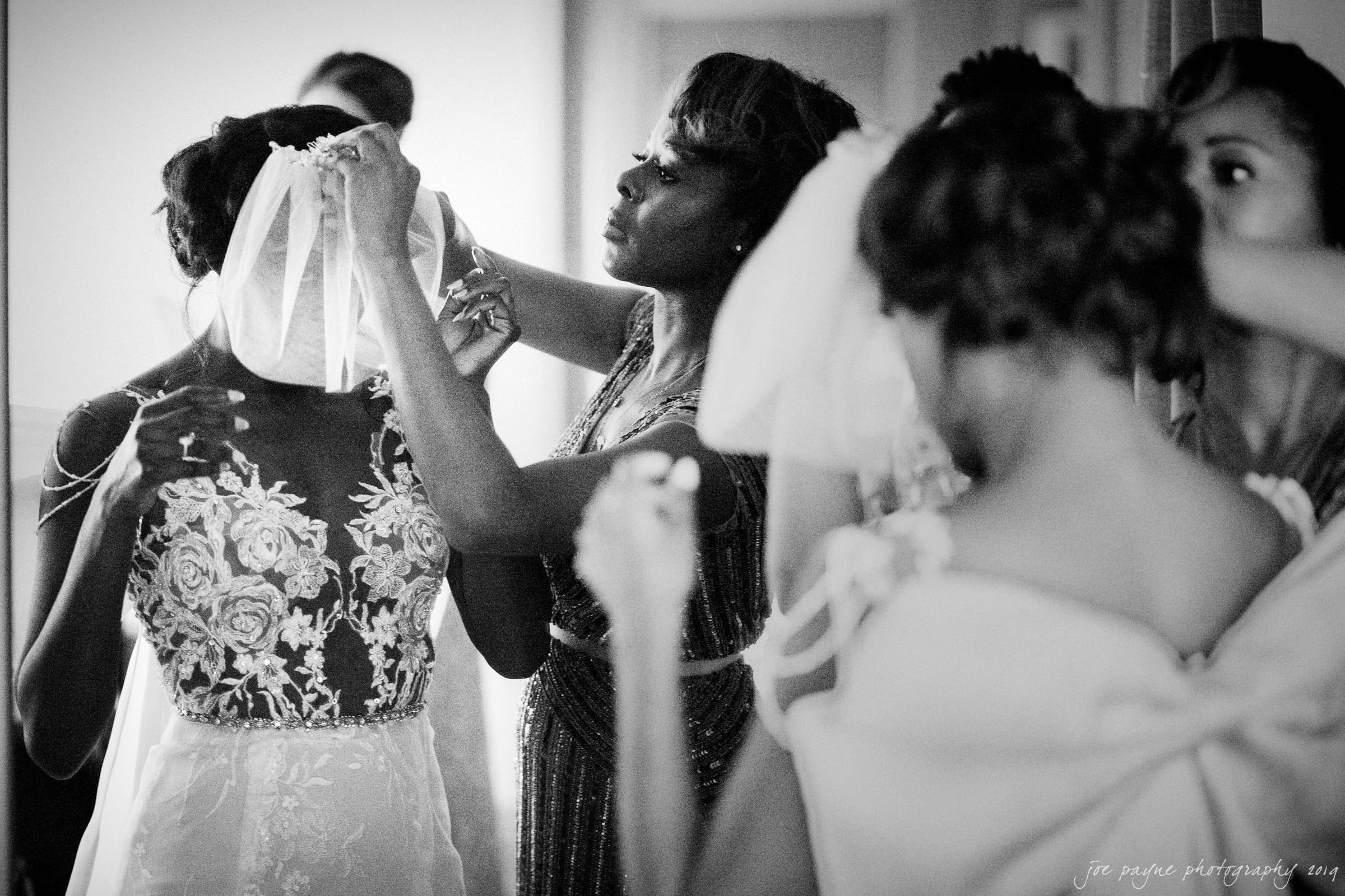21c museum hotel durham wedding - kortne & terry A black and white photo showing a bride having her veil adjusted by a woman. The bride wears a floral lace dress, and there are other bridesmaids nearby. The scene is filled with an atmosphere of anticipation and preparation.