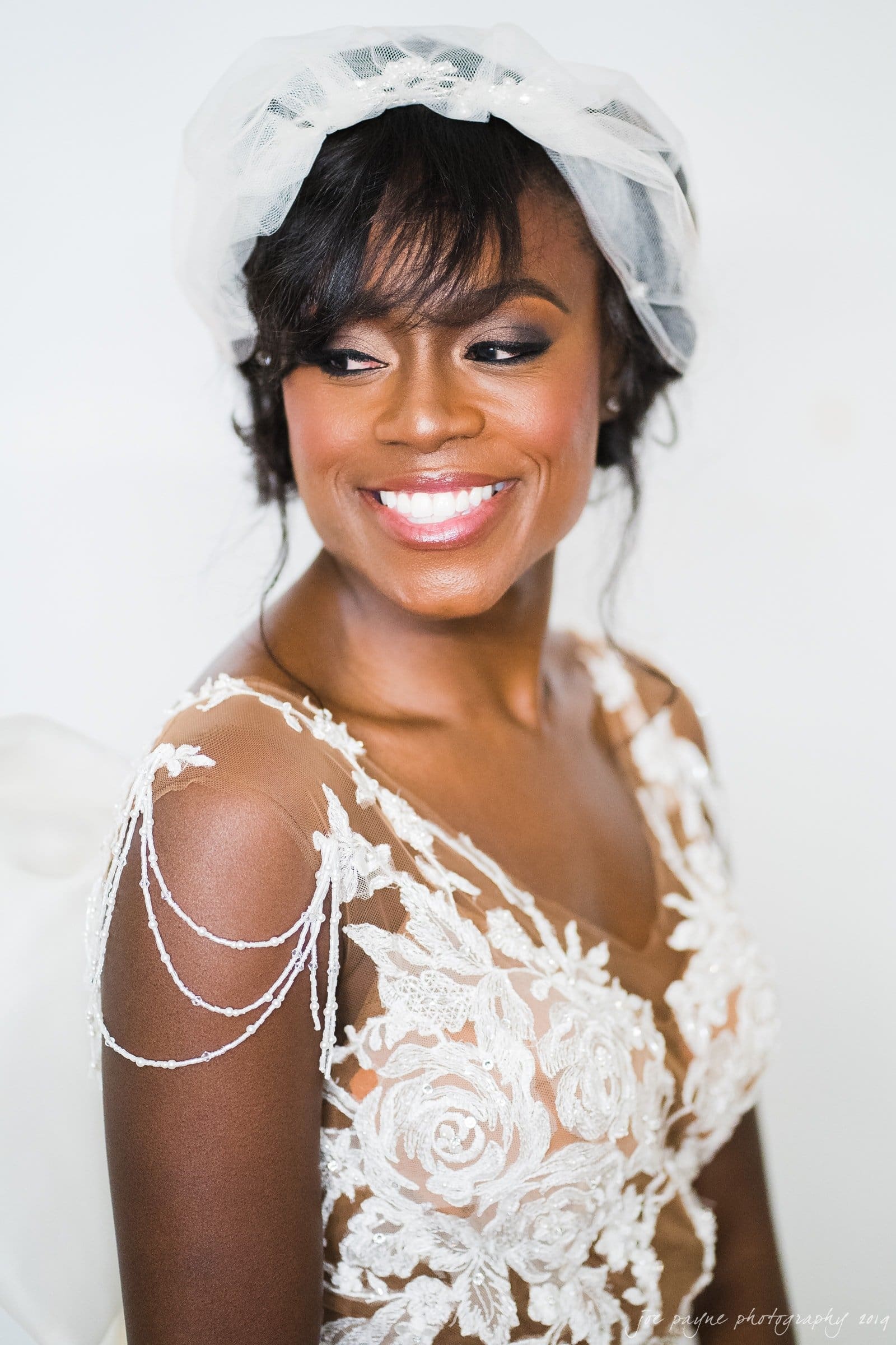 21c museum hotel durham wedding - kortne & terry A smiling bride wearing a delicate lace wedding dress with intricate floral patterns and beaded shoulder accents. She has a sheer veil over her dark hair, styled with soft curls, and is looking slightly to the side in a bright, softly lit setting.