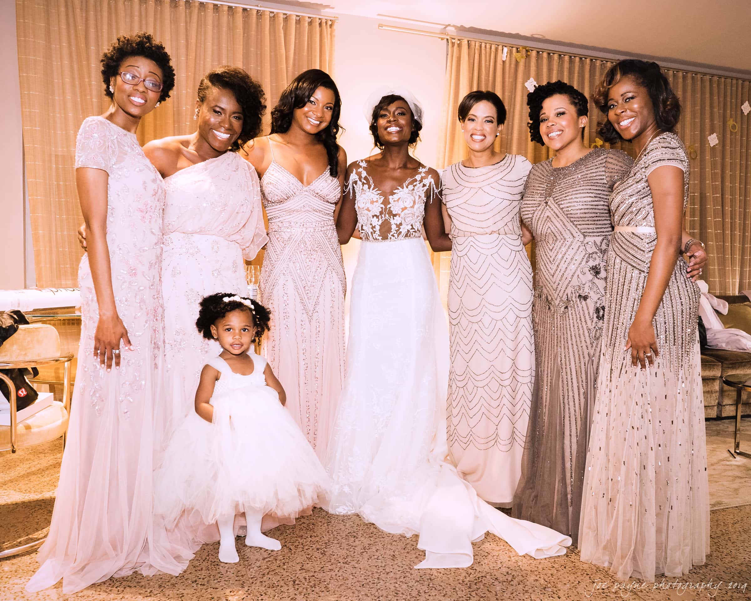 21c museum hotel durham wedding - kortne & terry A bride in a white gown stands with seven women dressed in various styles of elegant dresses, posing and smiling. A young girl in a white dress stands in front, smiling. Draped curtains are in the background.