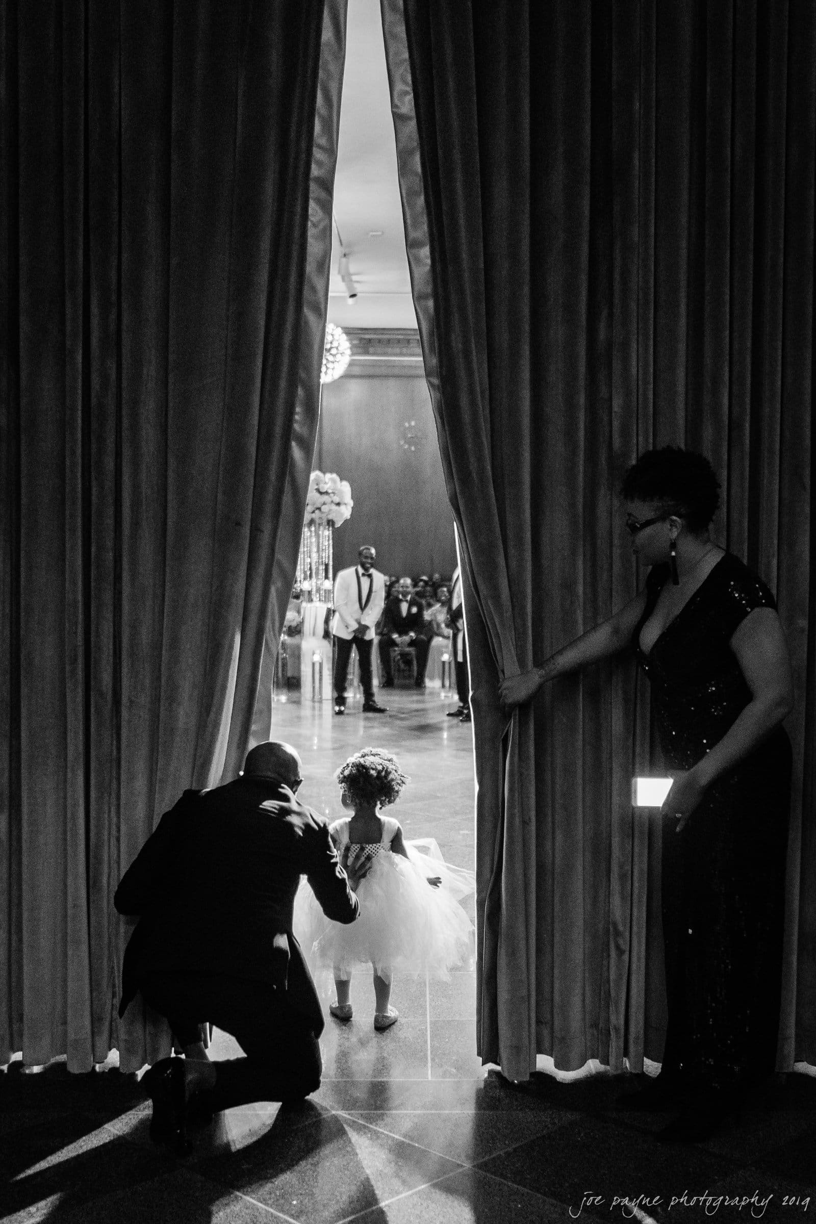 21c museum hotel durham wedding - kortne & terry A black and white photo showing a child in a white dress standing by a curtain with two adults. Theyre peeking into a room with balloons and people in the background.