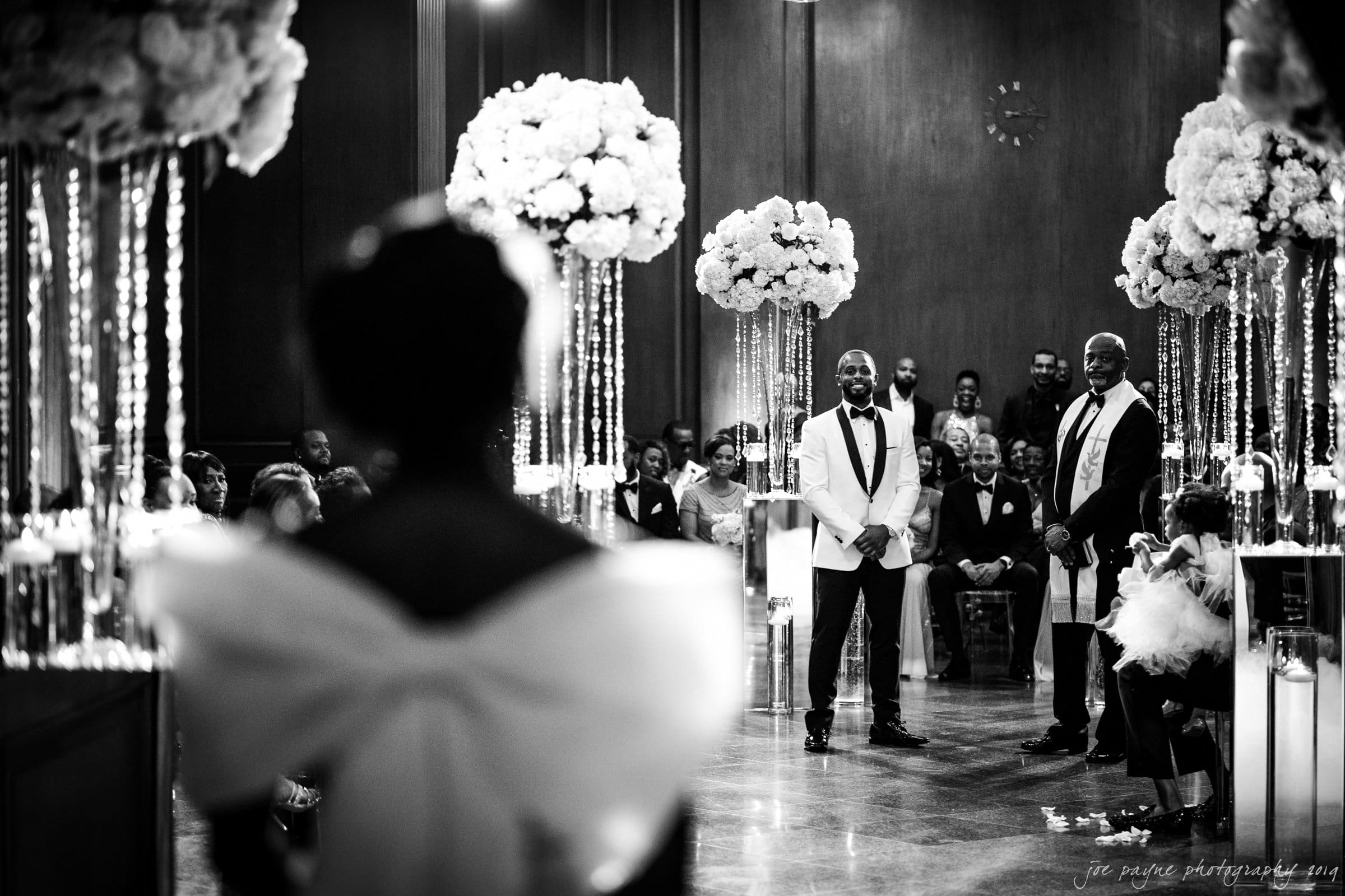 21c museum hotel durham wedding - kortne & terry A groom in a white tuxedo and the officiant stand at the altar, smiling as a bride in a white dress with a large bow approaches down the aisle. The venue is elegantly decorated with large floral arrangements and seated guests. Black and white photo.