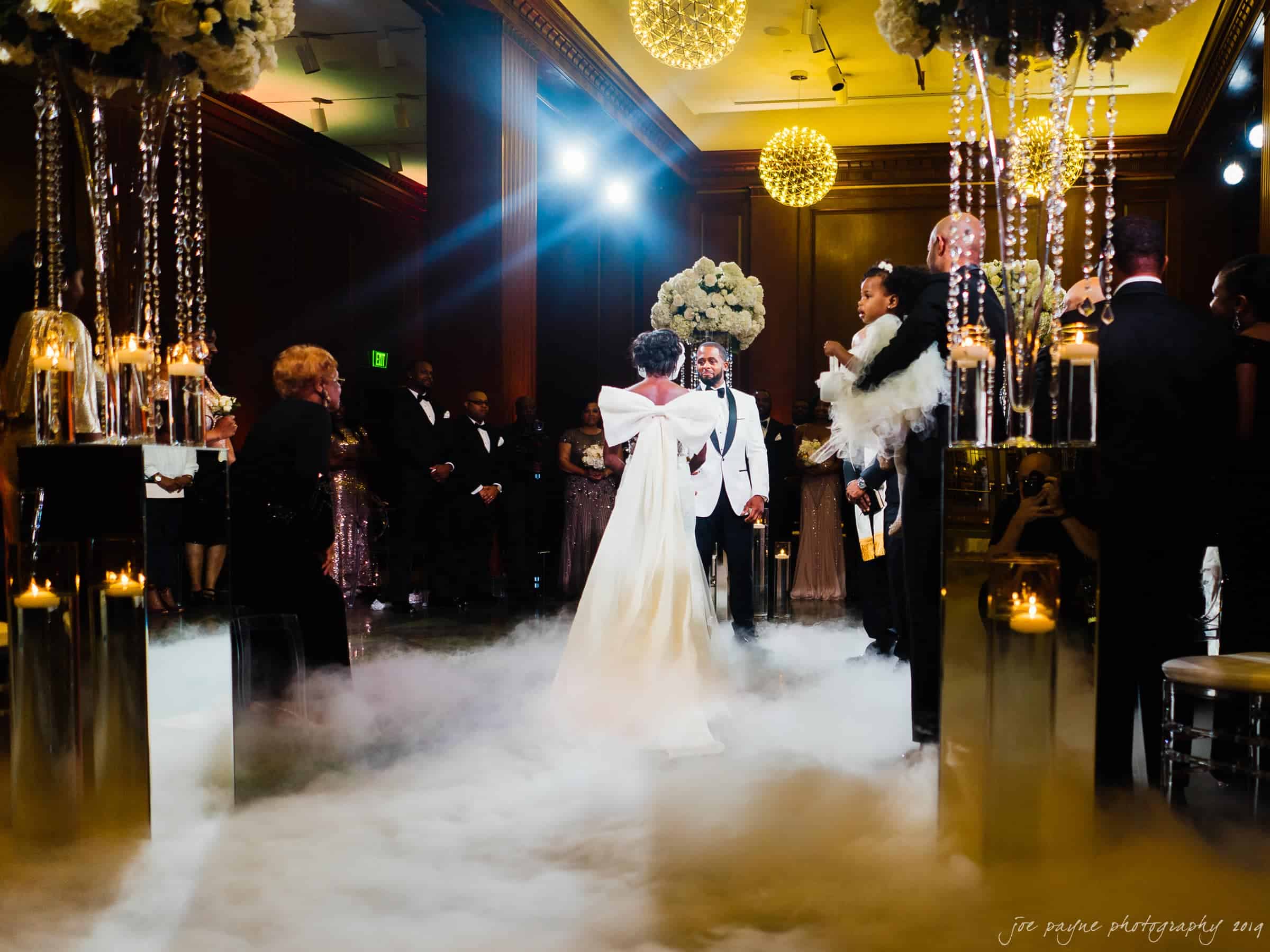 21c museum hotel durham wedding - kortne & terry A bride and groom share their first dance in an elegant venue. The floor is covered in a layer of mist, surrounded by guests in formal attire. The room features ornate chandeliers, decorative columns, and candlelit arrangements, creating a romantic ambiance.