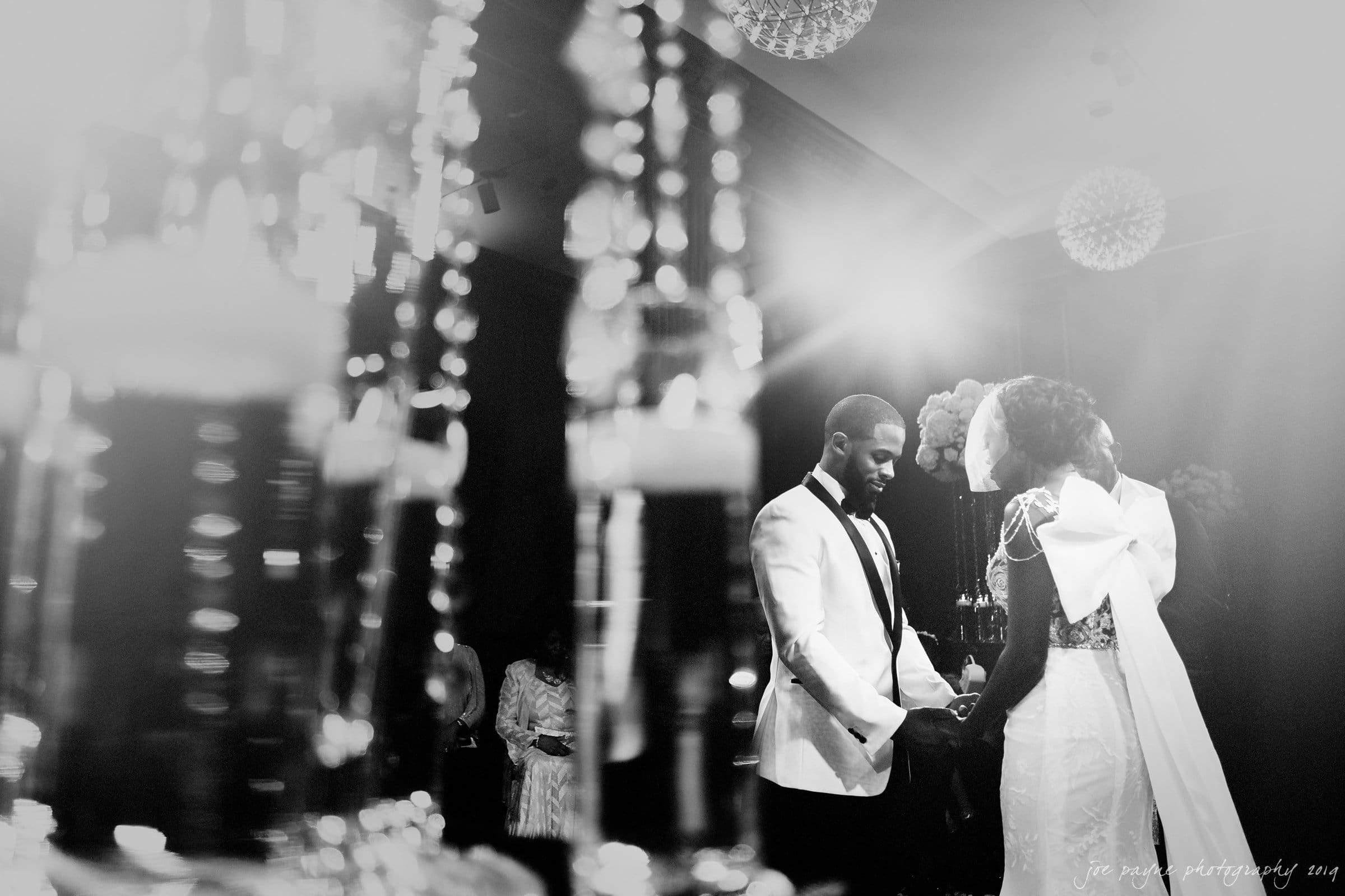 21c museum hotel durham wedding - kortne & terry A bride and groom stand facing each other, holding hands in a beautifully lit room. The groom wears a white jacket, while the bride wears a gown and veil. Decorative crystal elements and soft lighting enhance the elegant atmosphere.