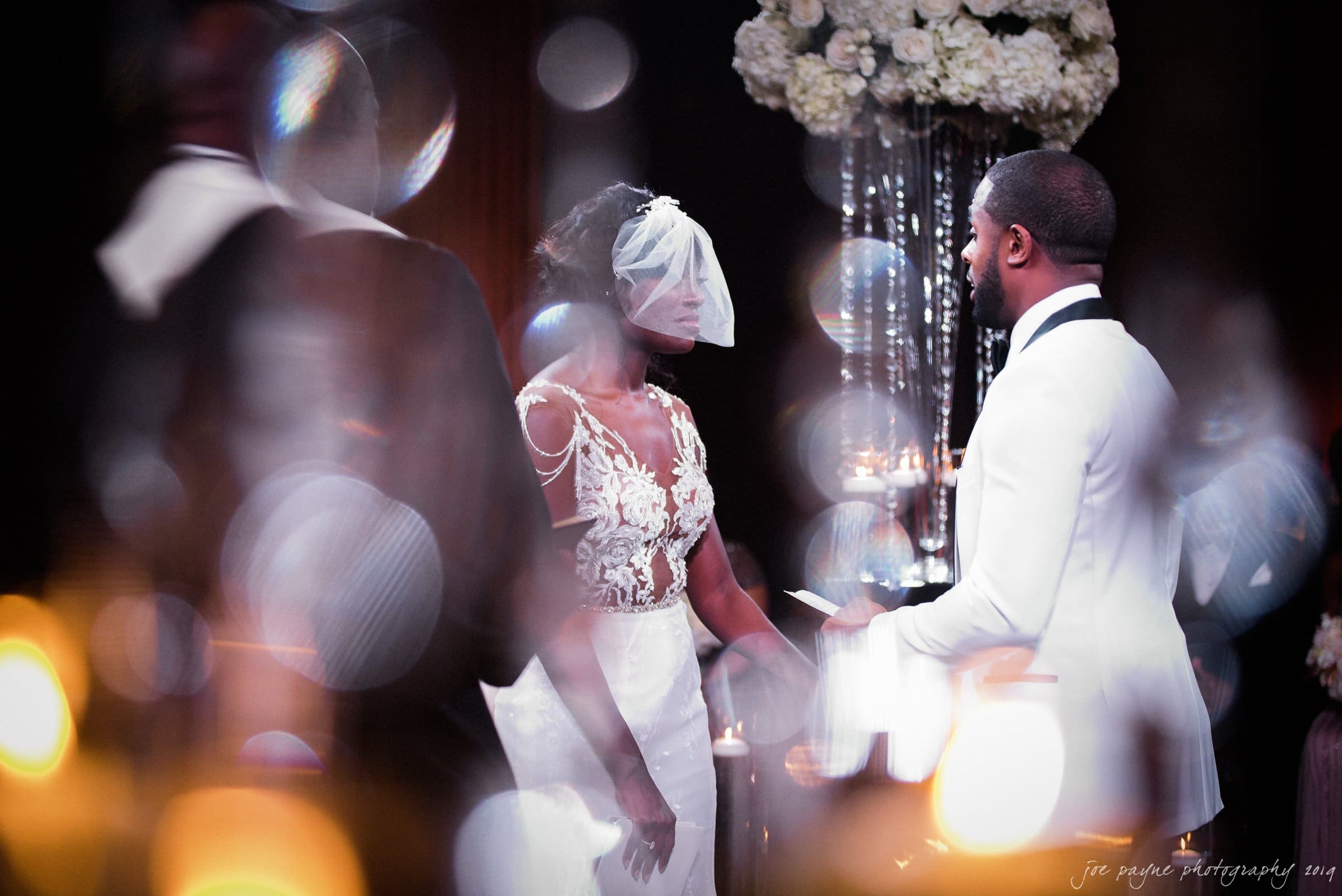 21c museum hotel durham wedding - kortne & terry A bride and groom face each other during a wedding ceremony. The bride wears a veil and a white gown with lace details, while the groom is in a white tuxedo. The scene is softly lit with blurred bokeh lights in the foreground.