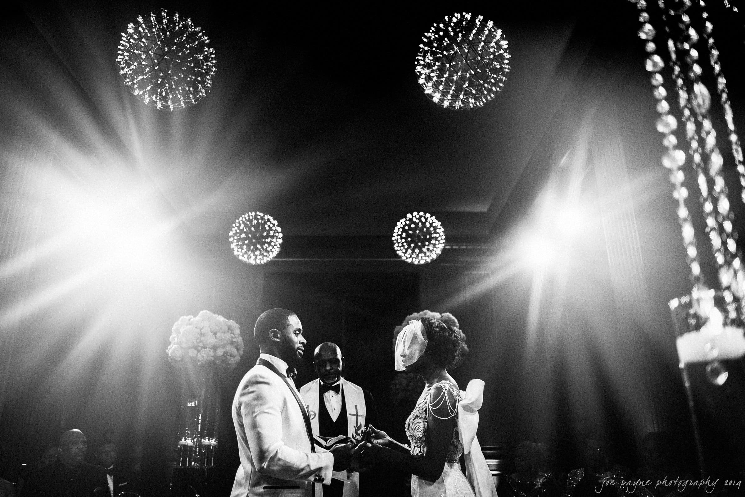21c museum hotel durham wedding - kortne & terry A black and white photo of a wedding ceremony. A bride and groom face each other, holding hands, with a smiling officiant between them. They are surrounded by elegant lighting and decorations, with guests visible in the background.