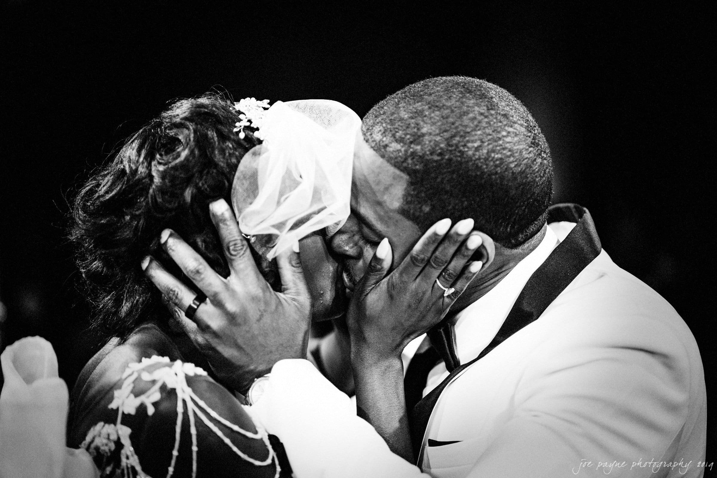 21c museum hotel durham wedding - kortne & terry A black and white photo of a couple sharing a kiss on their wedding day. The groom wears a white tuxedo, and the bride has a veil and floral hairpiece. They gently cradle each others faces, conveying a sense of intimacy and joy.