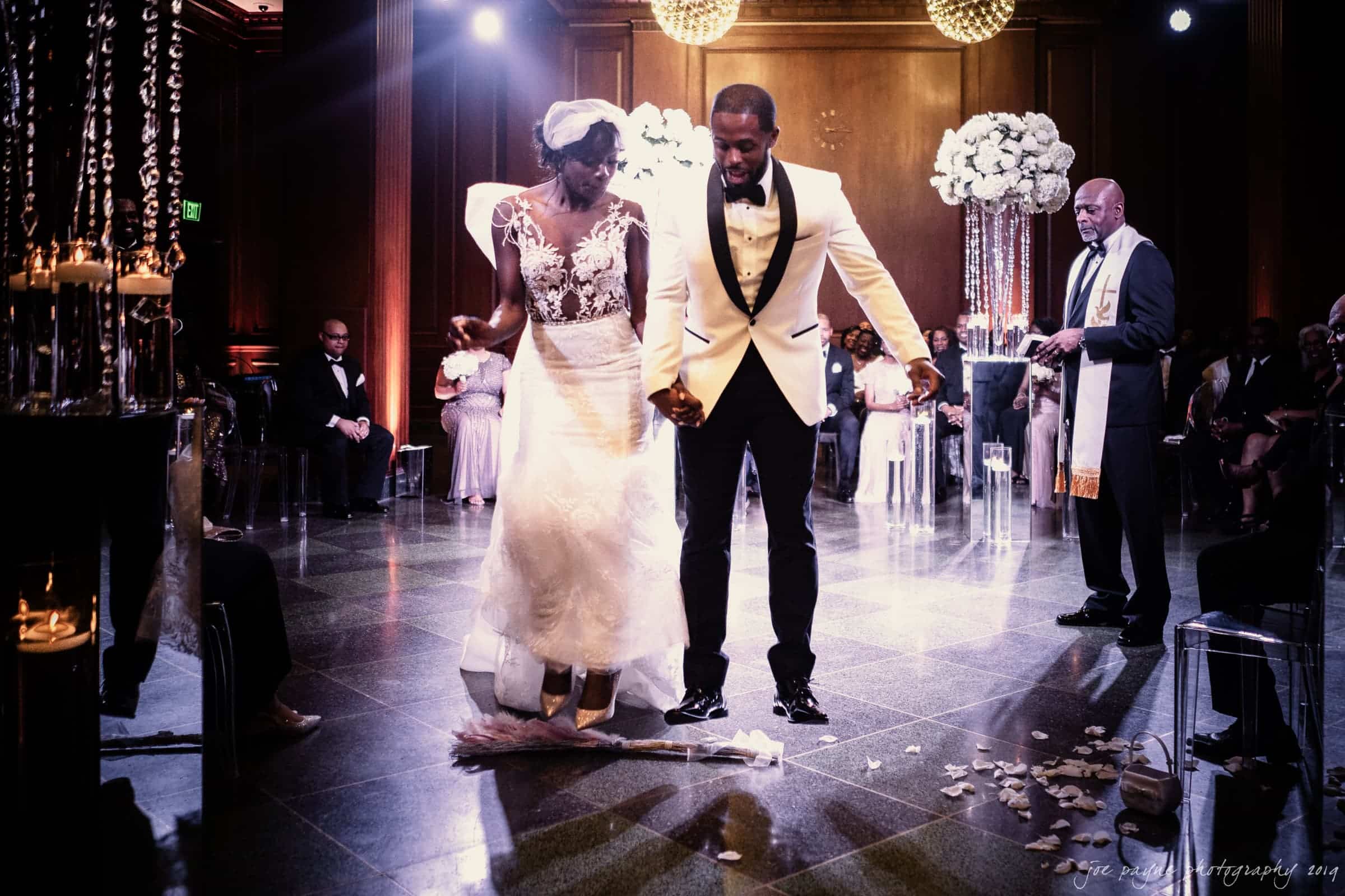 21c museum hotel durham wedding - kortne & terry A bride and groom in formal attire participate in a wedding ceremony, standing on a dimly lit, elegant venue floor with scattered petals. A decorated altar and a clergyman are visible in the background. Guests are seated around them.