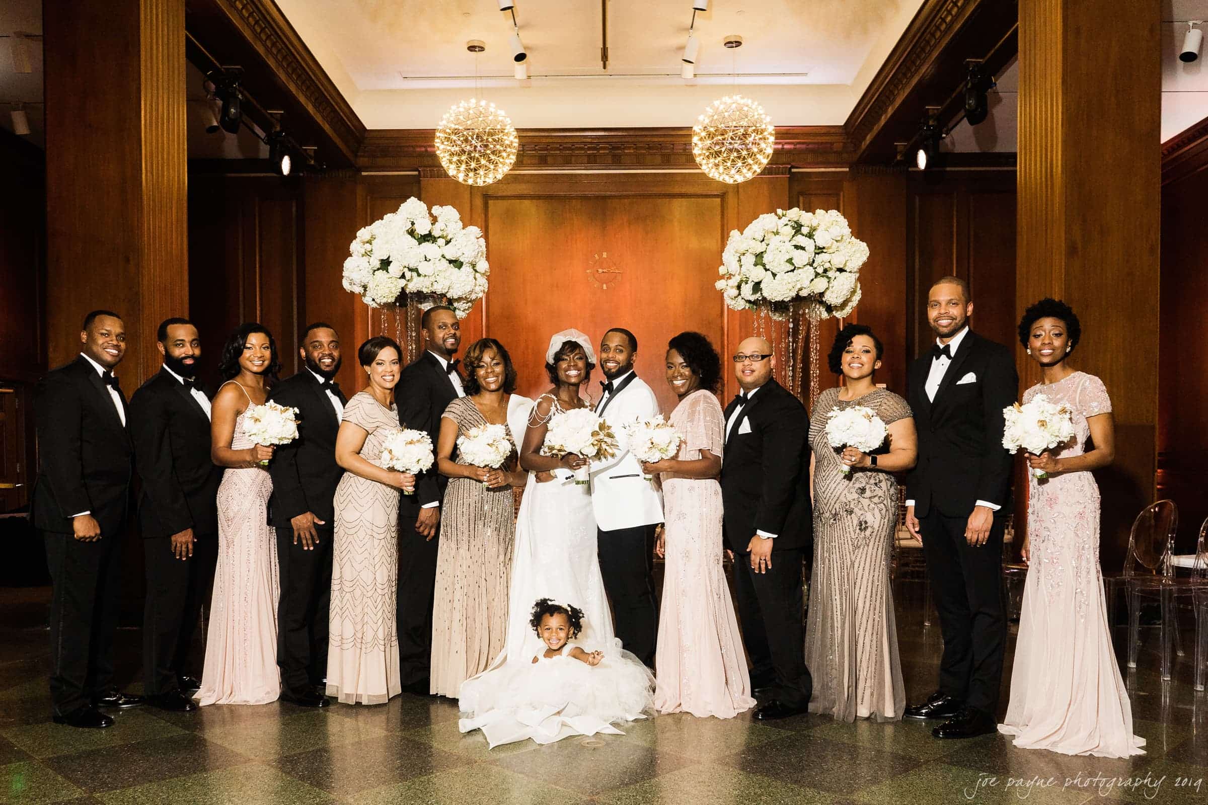 21c museum hotel durham wedding - kortne & terry A wedding party poses indoors. The bride and groom stand in the center, surrounded by bridesmaids in light pink dresses and groomsmen in black suits. A child sits at the front in a white dress. Decorations include flowers and elegant lighting.