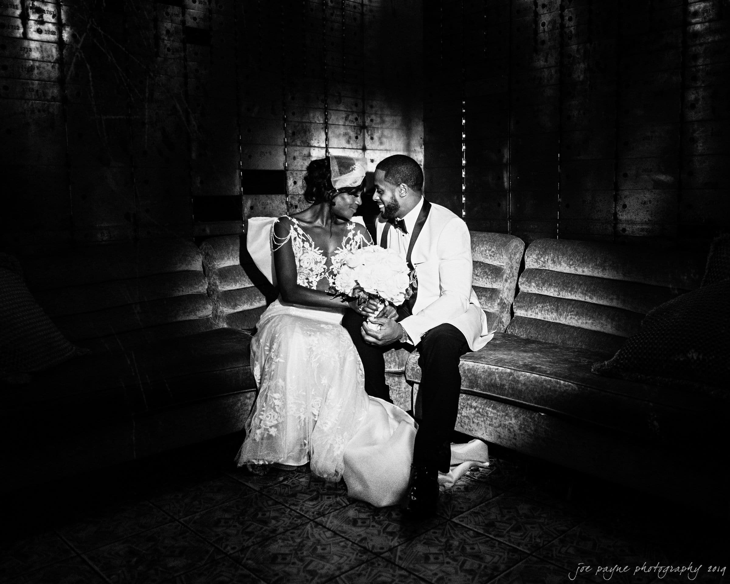 21c museum hotel durham wedding - kortne & terry A bride and groom sit closely on a vintage sofa in a dimly lit room. The bride holds a bouquet and wears a lace gown and veil. The groom is in a white tuxedo. They share an intimate gaze, highlighting the romantic atmosphere.