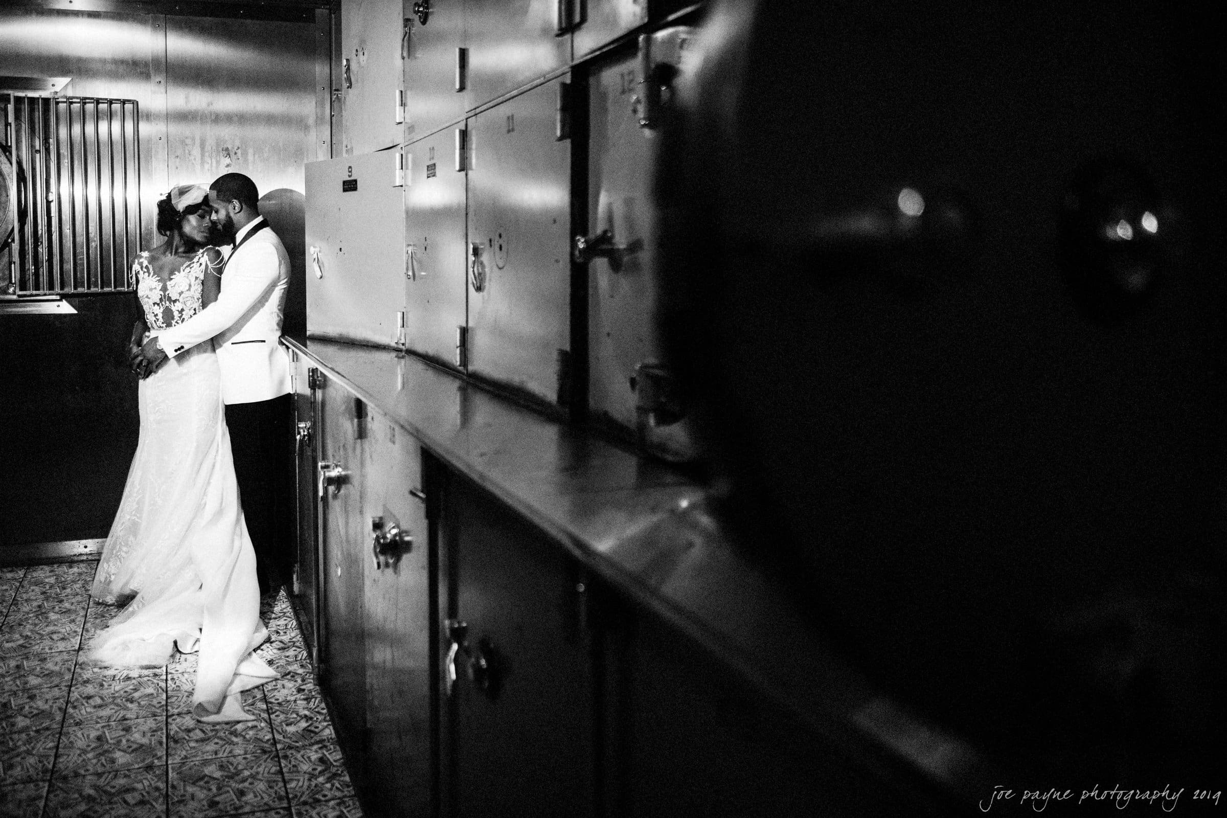 21c museum hotel durham wedding - kortne & terry A bride and groom share a romantic embrace in an industrial setting. She wears a long white gown with lace details, while he wears a white suit jacket. The dimly lit environment adds a dramatic, intimate atmosphere to the photograph.