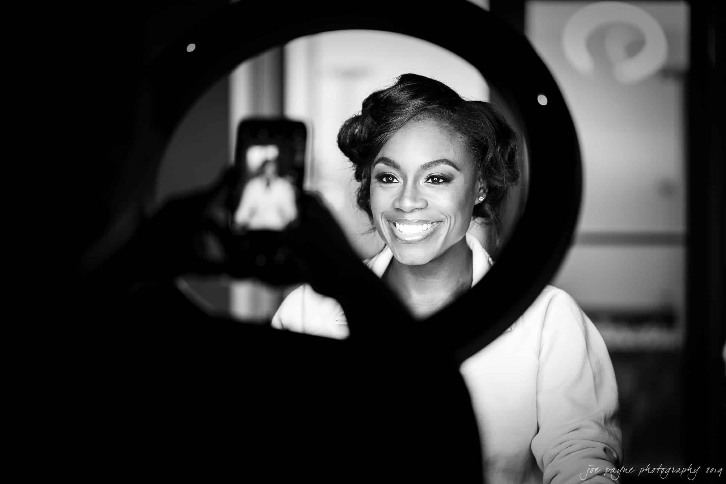 21c museum hotel durham wedding - kortne & terry A woman smiles while being photographed through a circular light. The scene is captured in black and white, highlighting her joyful expression. She is looking at a phone that someone is using to take her picture.